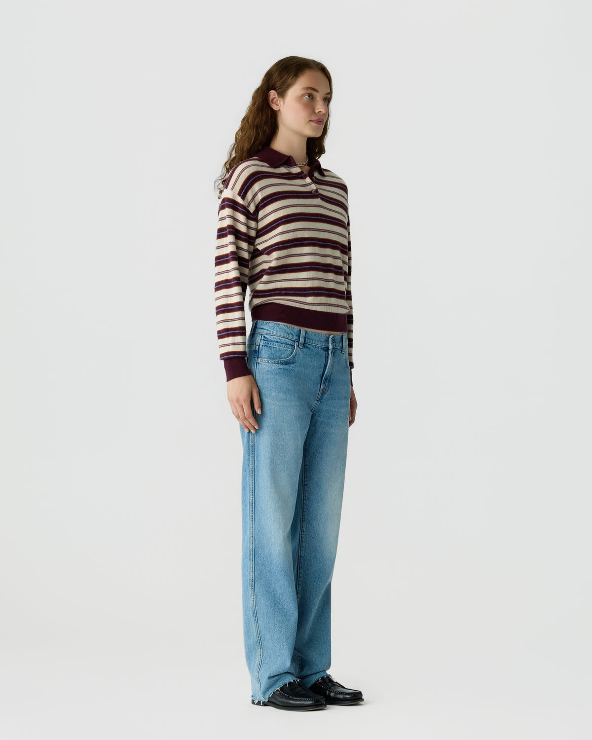 Woman standing in a side profile wearing a striped long-sleeve sweater with a collar, light blue wide-leg jeans, and black shoes against a plain white background.