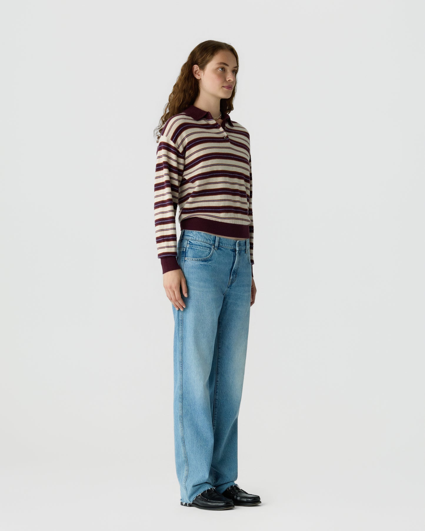 Woman standing in a side profile wearing a striped long-sleeve sweater with a collar, light blue wide-leg jeans, and black shoes against a plain white background.