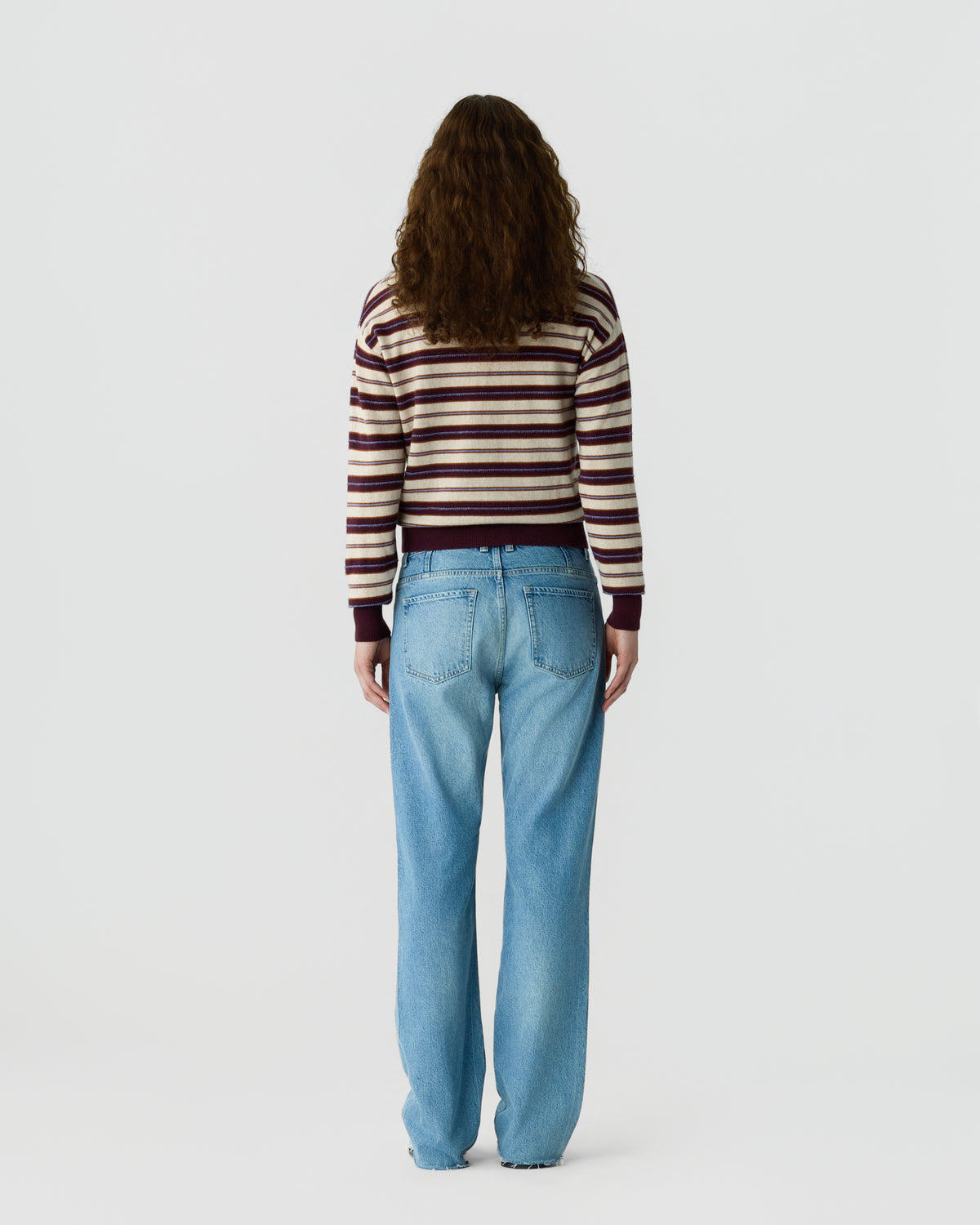 Person with long curly hair wearing a striped sweater and light blue jeans standing facing away against a plain white background