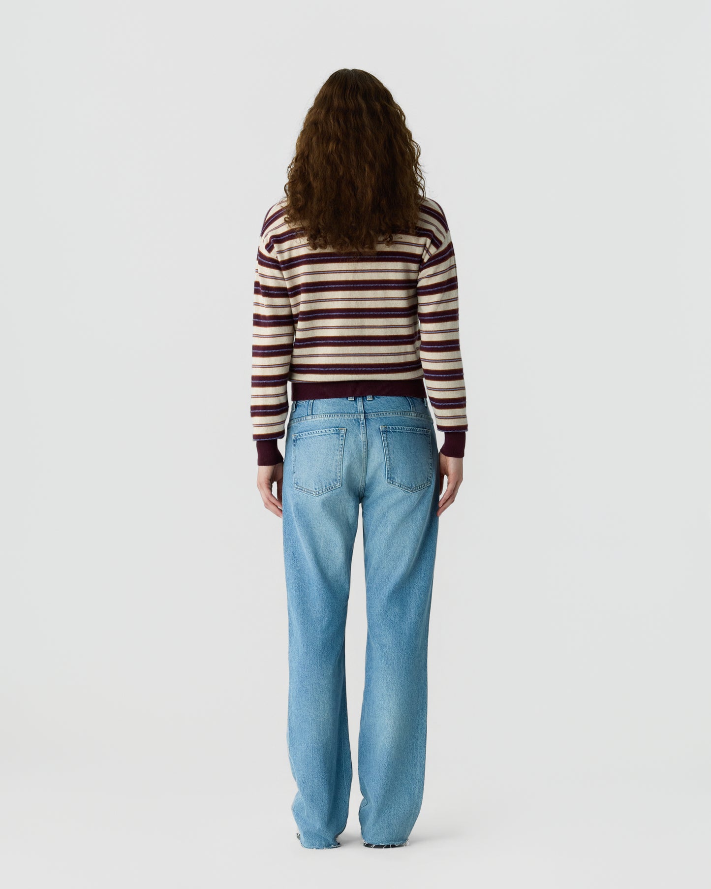 Person with long curly hair wearing a striped sweater and light blue jeans standing facing away against a plain white background