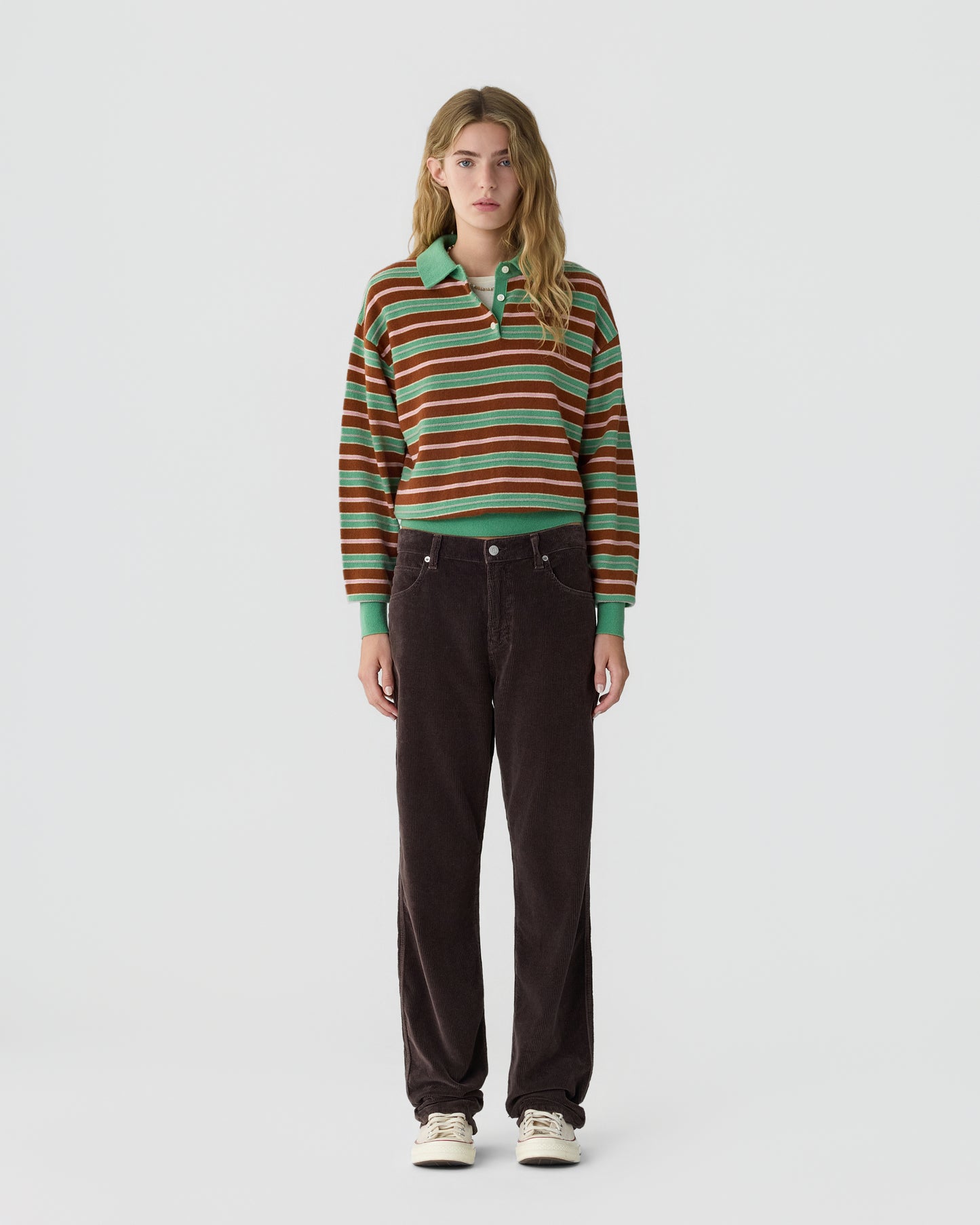 Young woman standing against a plain white background wearing a green, brown, and white striped long-sleeve polo shirt with dark brown pants and white sneakers.