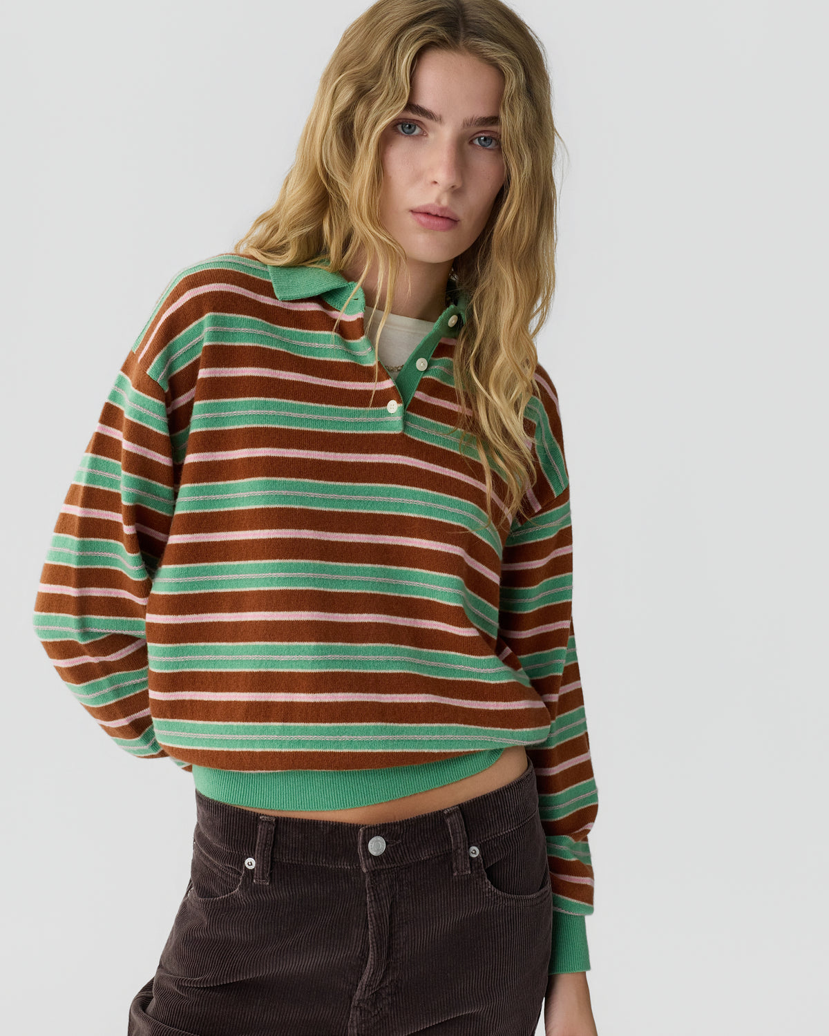 Young woman with long wavy blonde hair wearing a green and brown striped long-sleeve polo shirt and dark brown corduroy pants against a plain gray background.