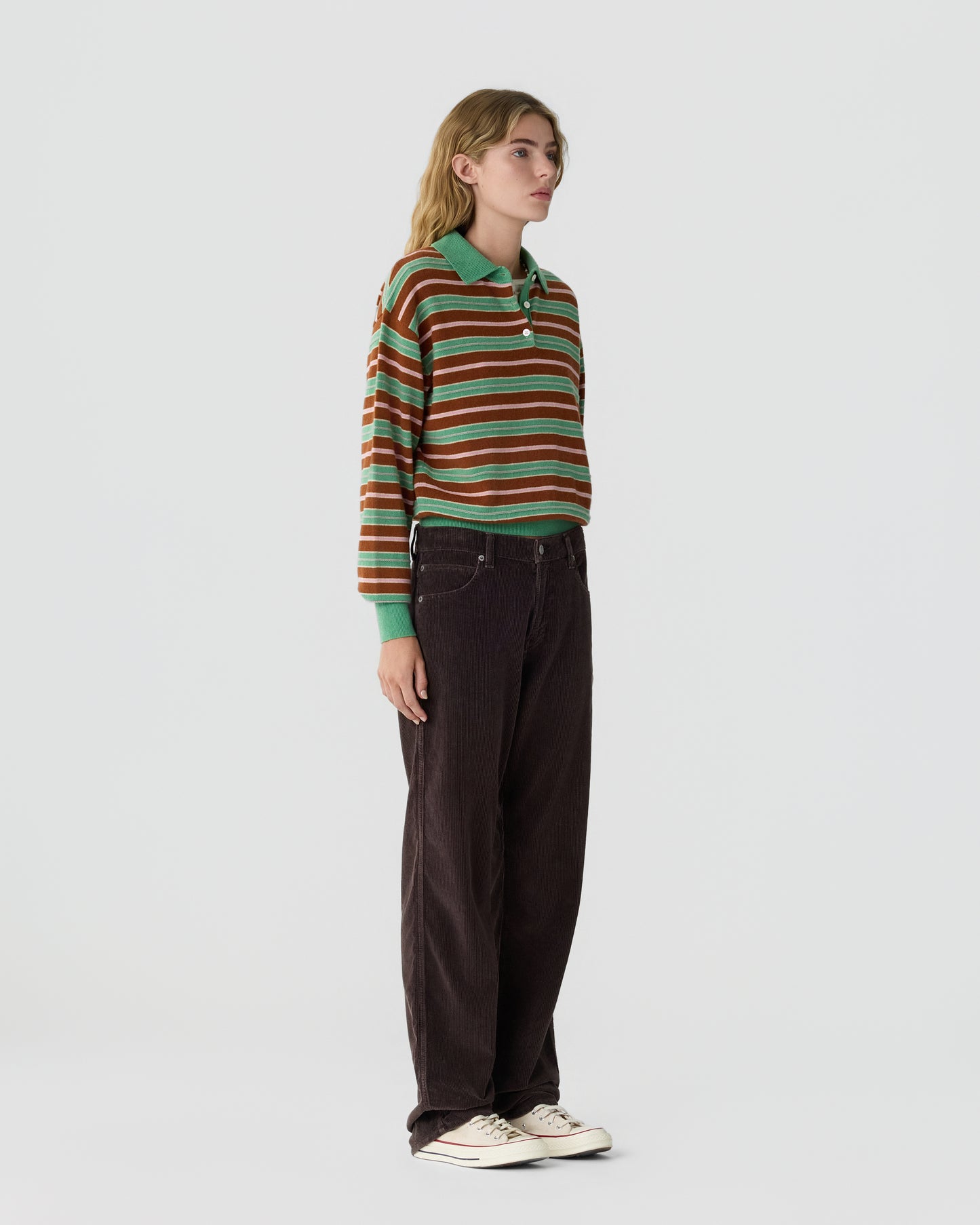 Young woman standing wearing a striped long-sleeve polo shirt with green, brown, and white stripes paired with dark brown pants and white sneakers on a plain light background.