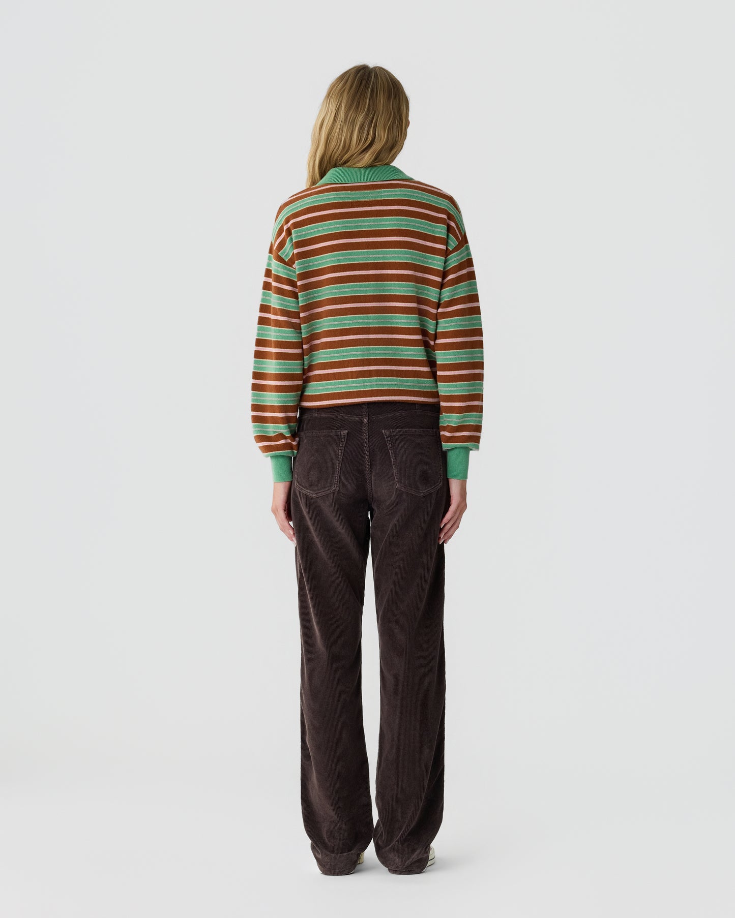 Person standing facing away from the camera wearing a green, brown, and white striped long-sleeve sweater and dark brown pants against a plain light background.