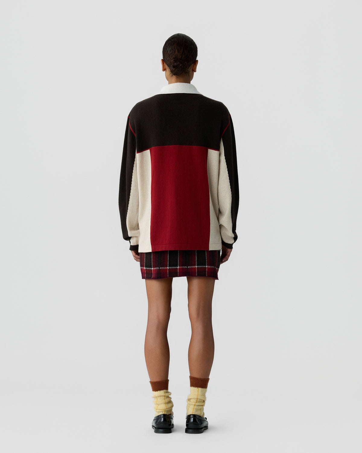 Person wearing a color-blocked sweater with black, cream, and red sections paired with a red and black plaid skirt, standing against a plain gray background.