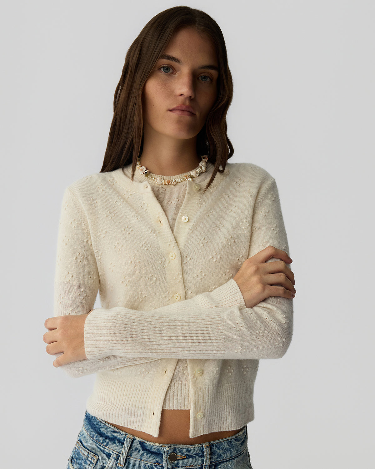 Young woman wearing a cream-colored button-up cardigan with a delicate textured pattern over a matching top, paired with light wash denim jeans against a plain background.