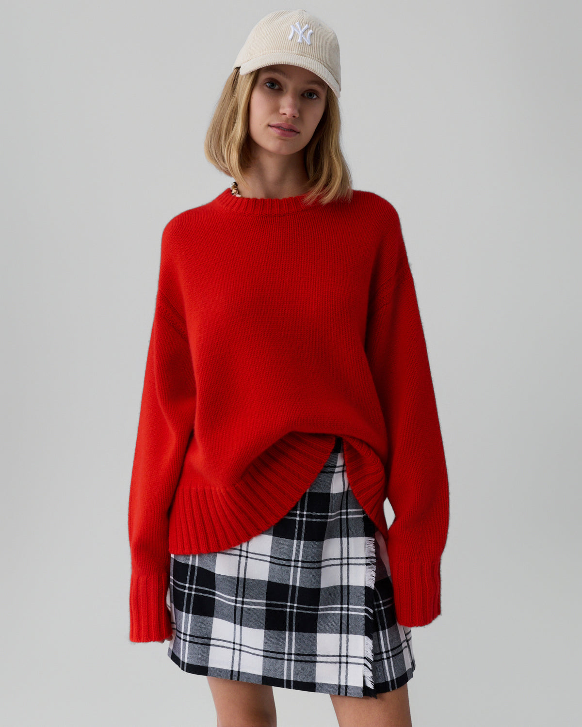 Young woman wearing an oversized bright red knit sweater with ribbed cuffs and hem, paired with a black and white plaid skirt and a beige baseball cap against a plain gray background.