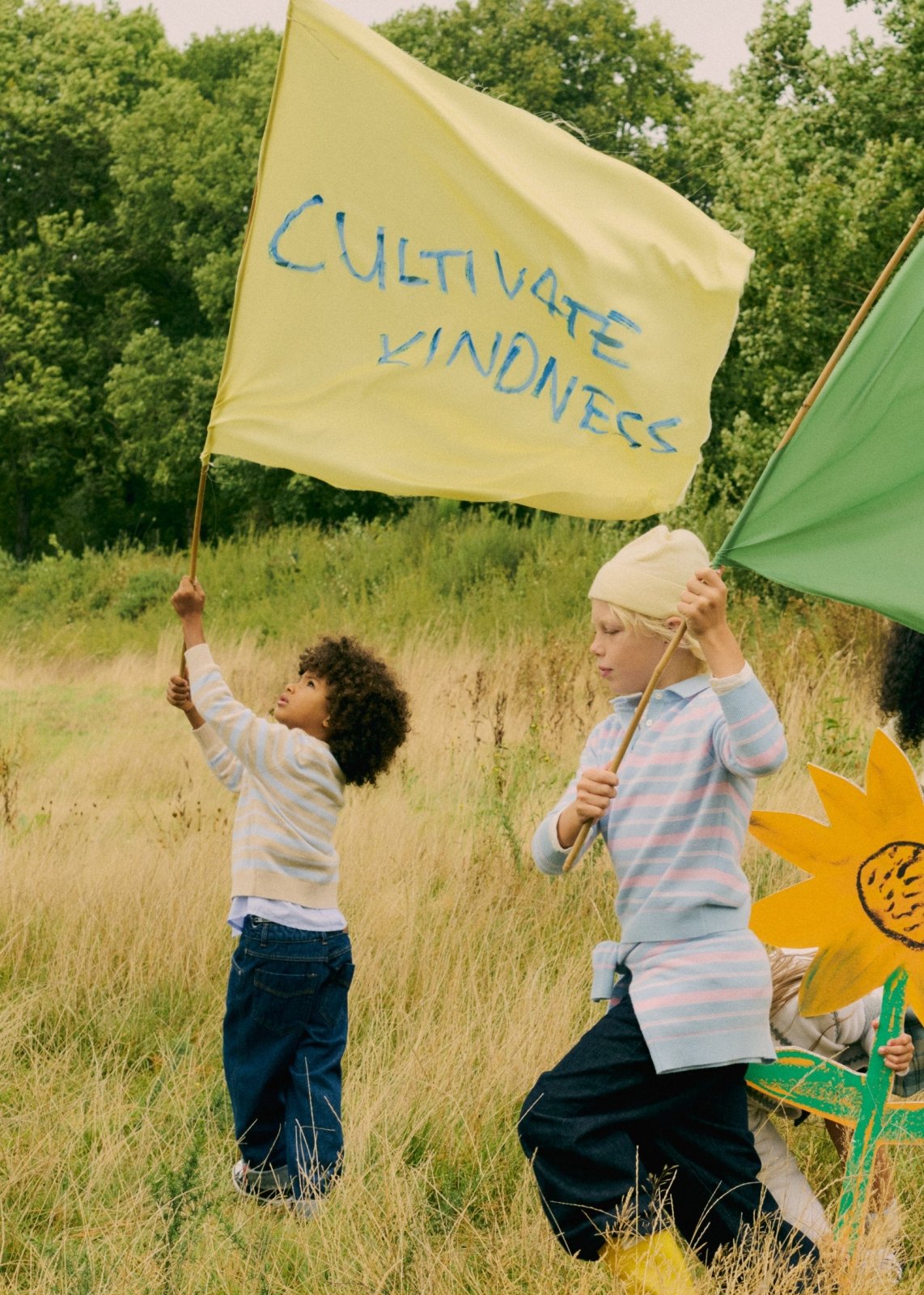 Children outdoors in casual Bonpoint clothing holding colorful flags and a large sunflower prop in grassy field.