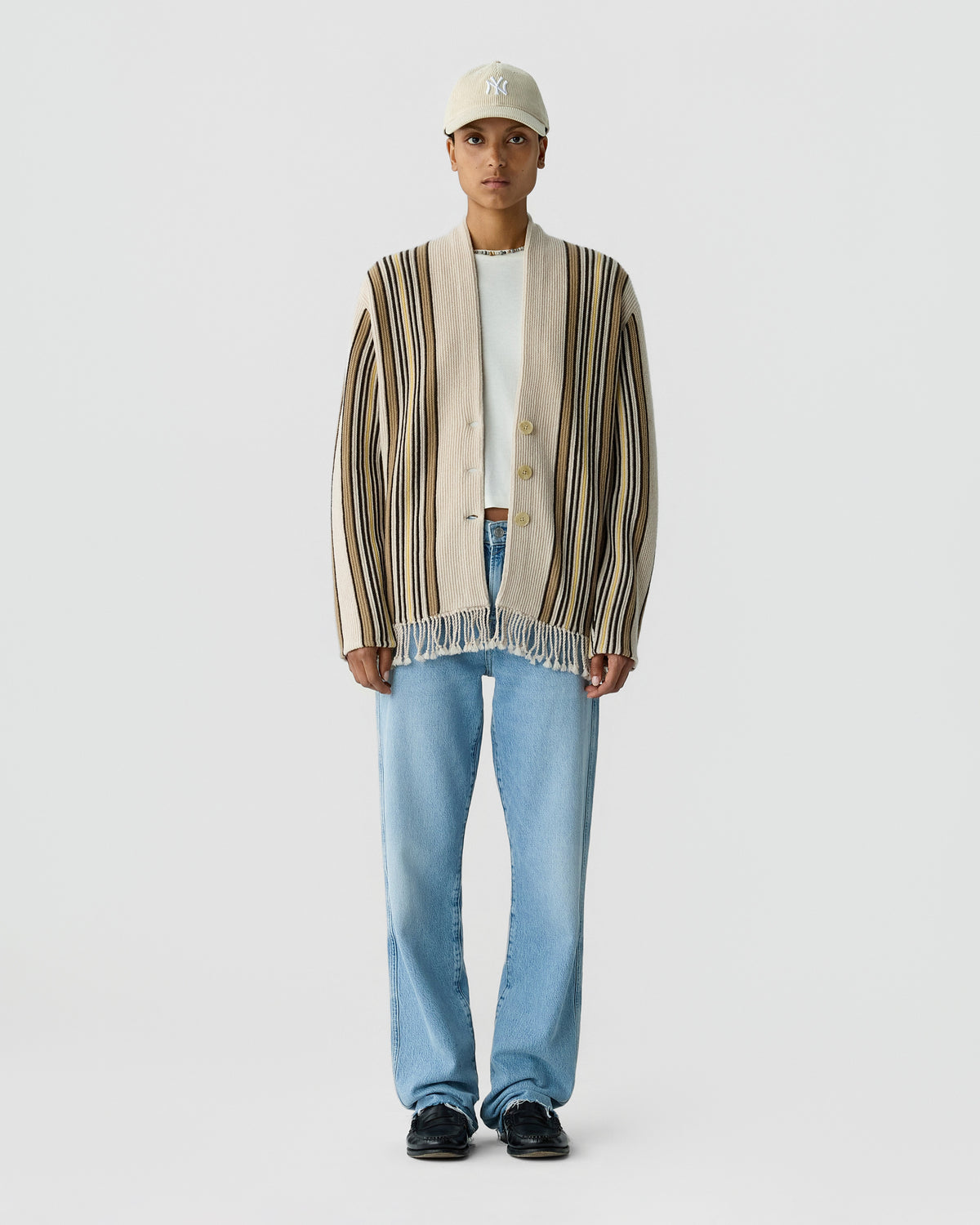 Person wearing a striped button front cardigan over a white shirt with blue jeans a light cap and black shoes against a plain gray background