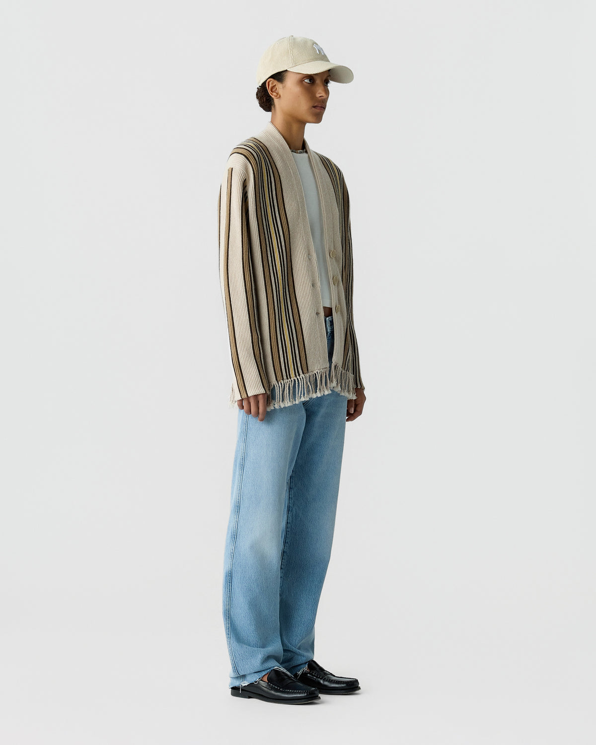 Person wearing a striped fringed cardigan over a light shirt with loose blue jeans a baseball cap and black loafers against a pale background