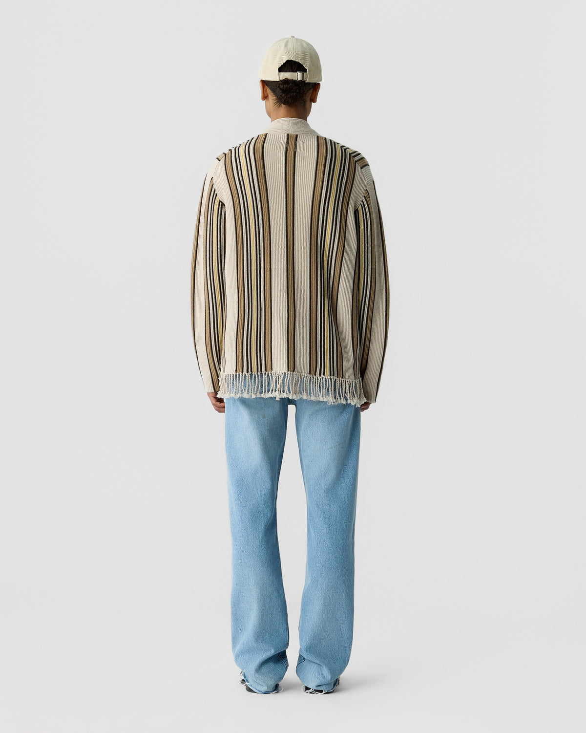 Back view of a model wearing a striped knit cardigan with fringe trim, light blue jeans, and a cream baseball cap against a pale background