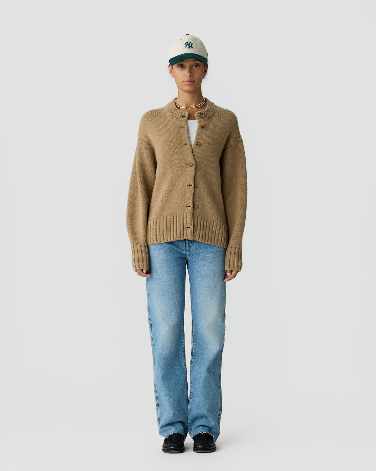 Person wearing a beige cashmere cardigan over a white top, light blue jeans, black shoes, and a white and green baseball cap, standing against a plain background.