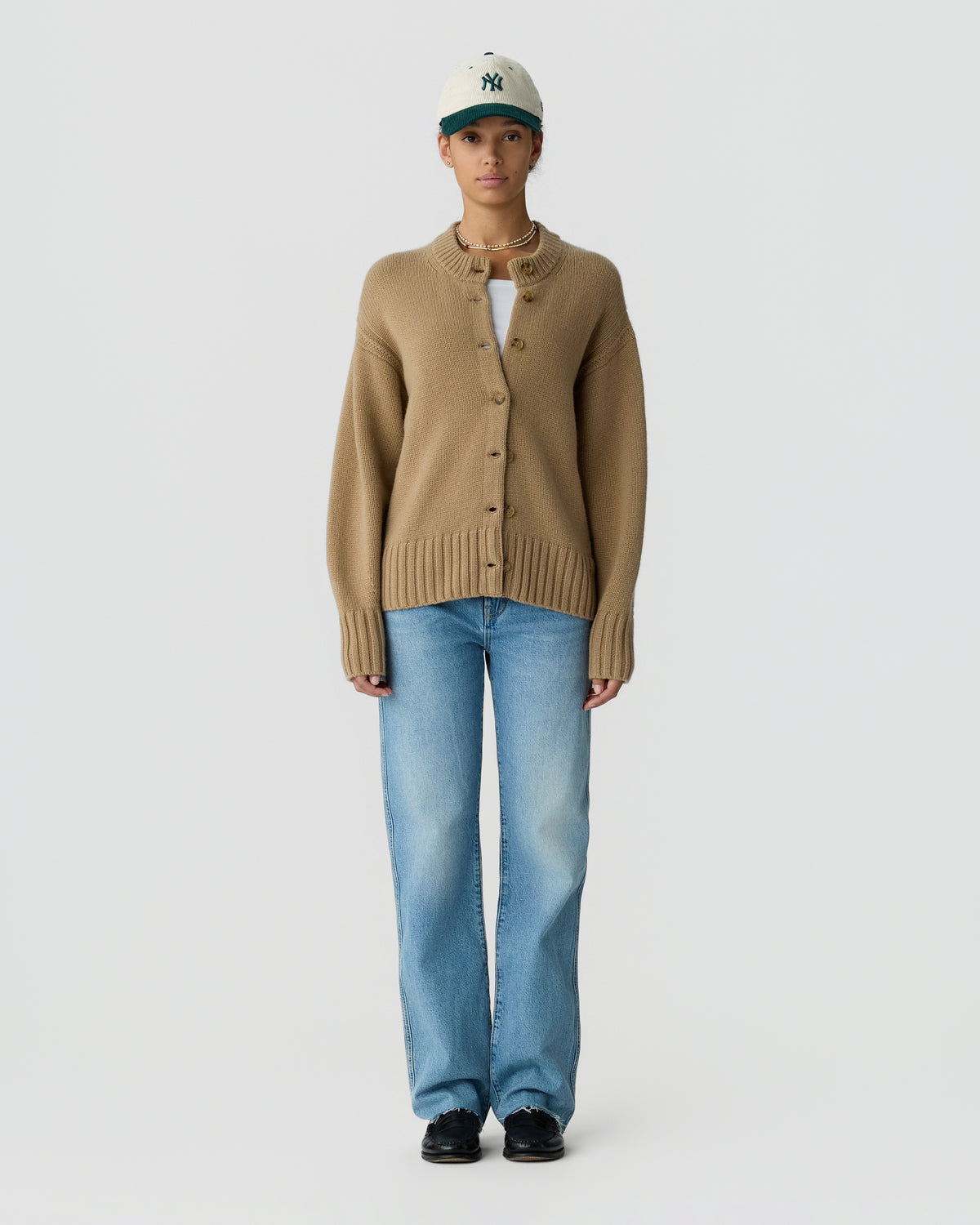 Model wearing a brown button front cardigan, light blue jeans, black shoes, layered necklaces, and a gray green cap against a plain light background