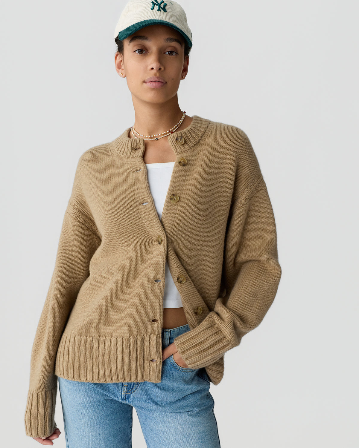 Model wearing a beige button front cardigan over a white top, blue jeans, layered necklaces, and a baseball cap against a light gray background