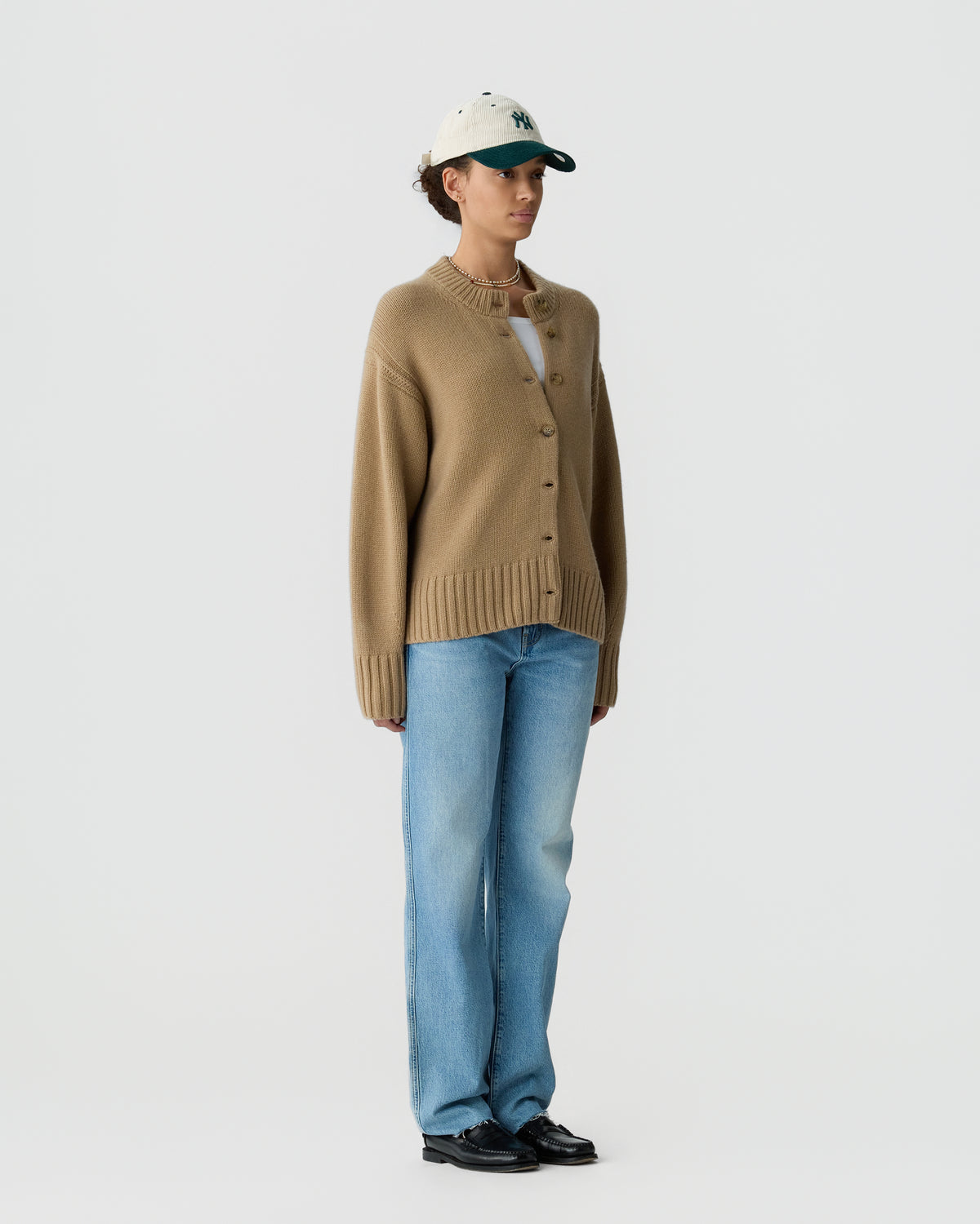 Woman wearing a beige button-up cashmere cardigan over a white top, light blue straight-leg jeans, black shoes, and a white baseball cap against a plain white background.