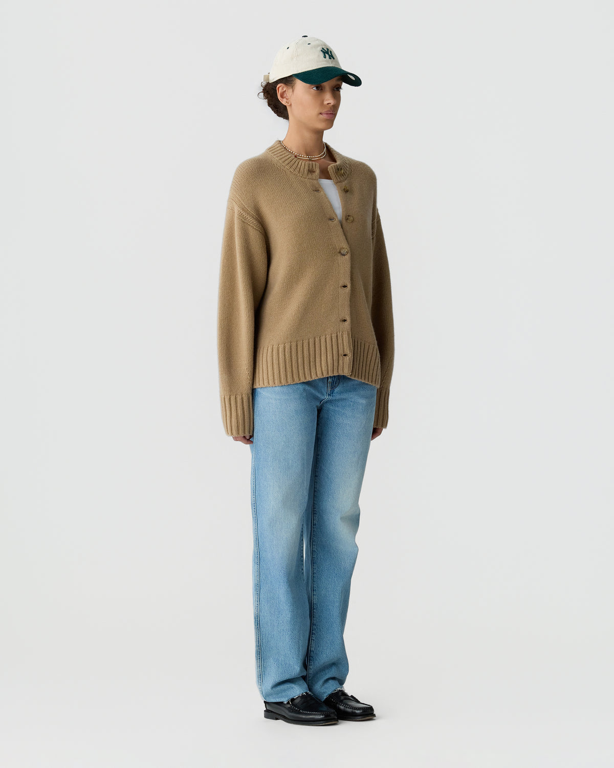 Model wearing a brown button front cardigan with blue jeans, black shoes, and a light cap against a plain gray studio background.