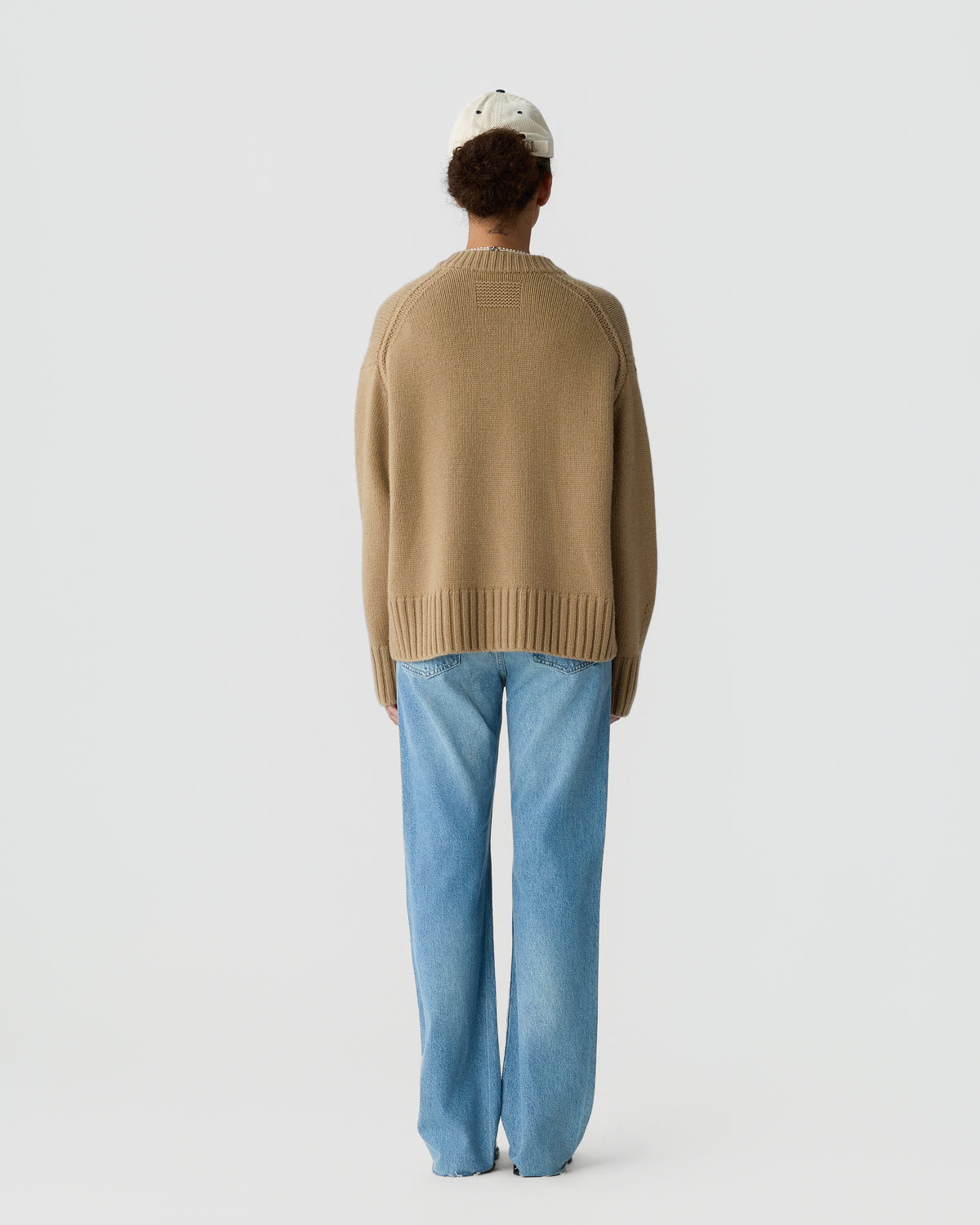 Person wearing a beige oversized sweater and light blue wide-leg jeans, standing with their back to the camera against a plain background.