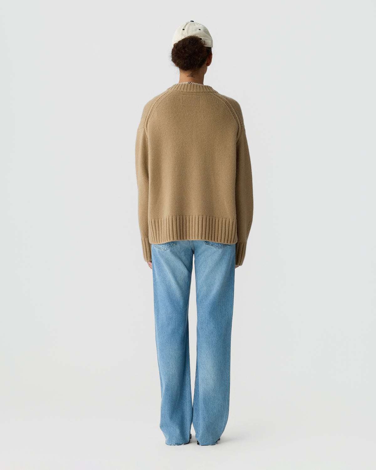 Person viewed from behind wearing a brown knit sweater with light blue jeans and a white cap against a pale background