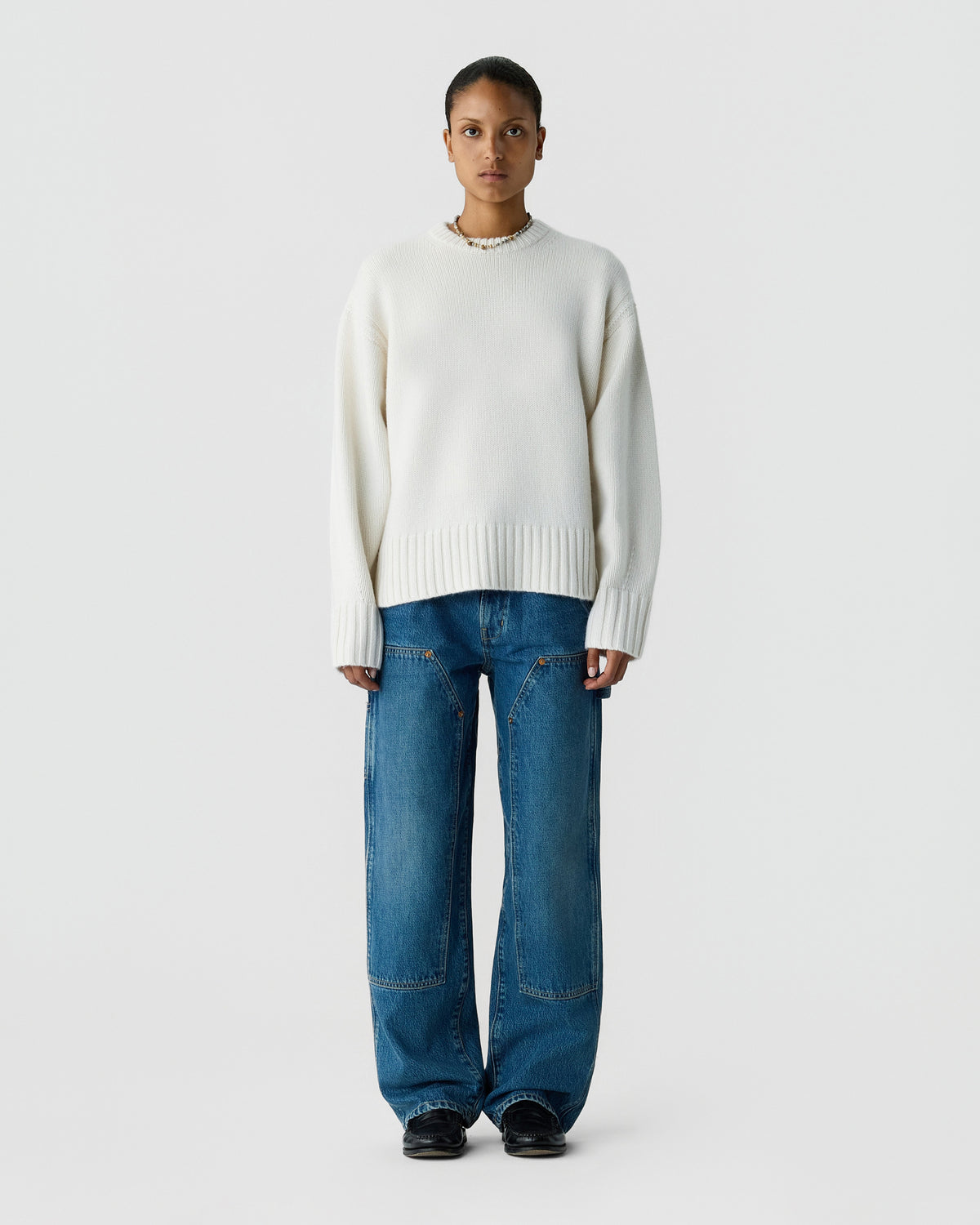 Model wearing a white crew neck sweater with ribbed cuffs and hem plus relaxed blue jeans and black shoes against a light gray background