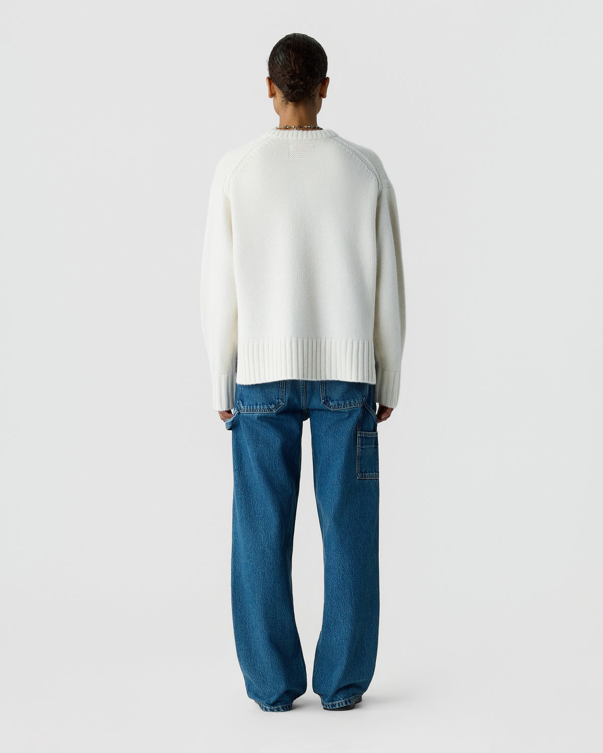 Back view of a person wearing a cream knit sweater with ribbed cuffs and hem and relaxed blue jeans against a light gray background