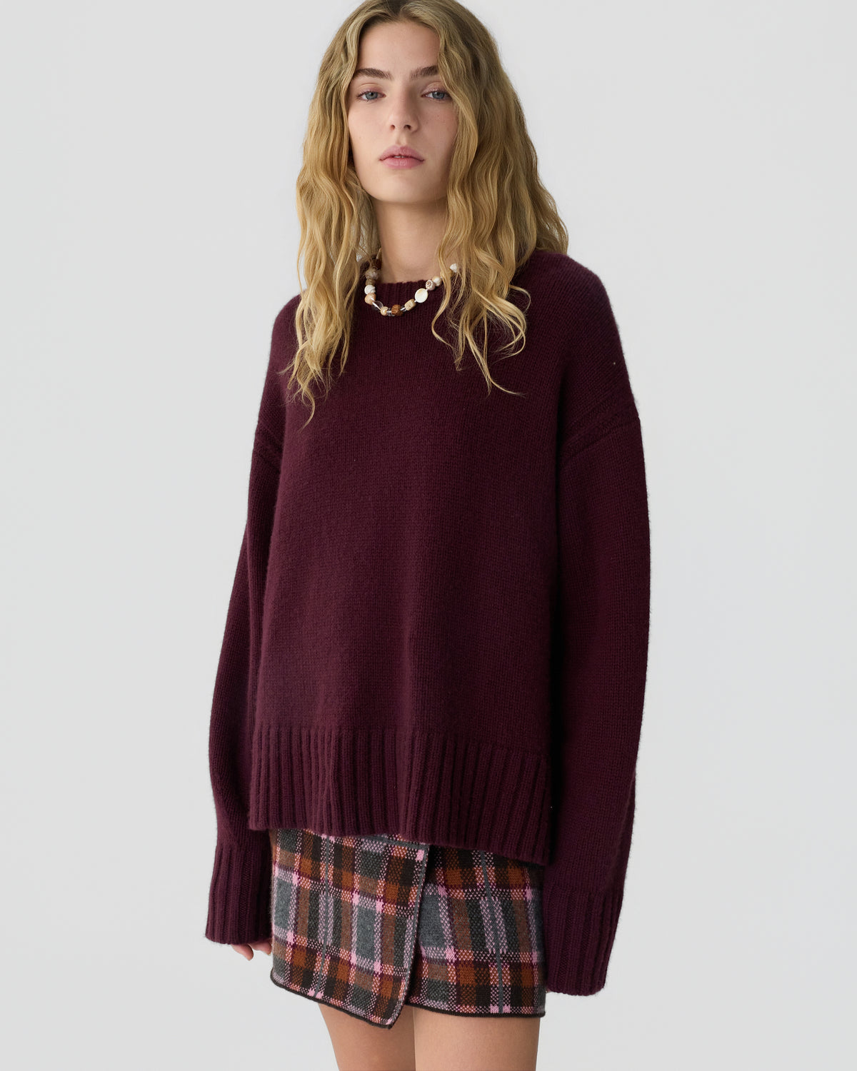 Young woman with long wavy blonde hair wearing a loose-fitting deep burgundy cashmere sweater and a plaid skirt with shades of brown, pink, and gray against a plain light background.