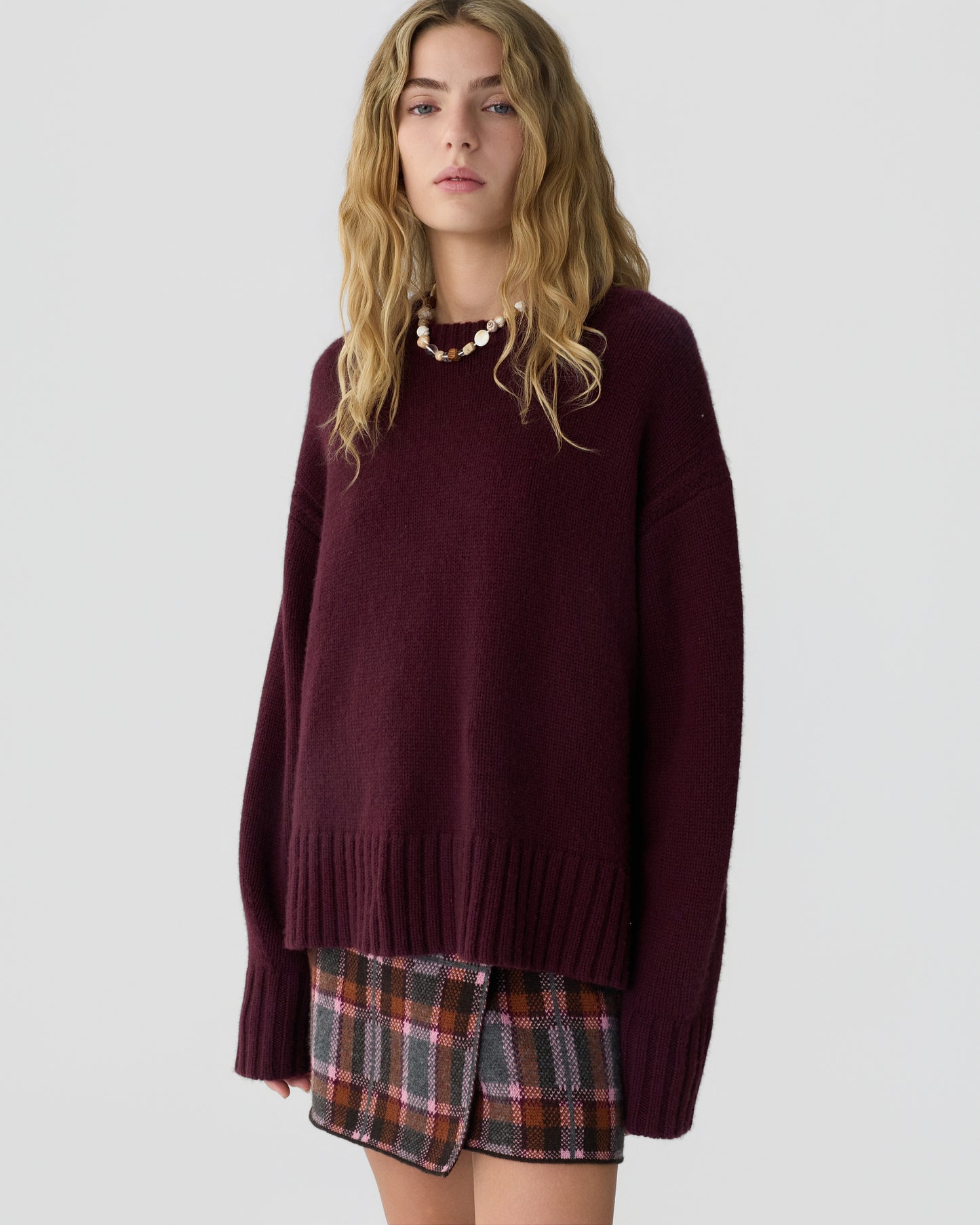 Young woman with long wavy blonde hair wearing a loose-fitting deep burgundy cashmere sweater and a plaid skirt with shades of brown, pink, and gray against a plain light background.