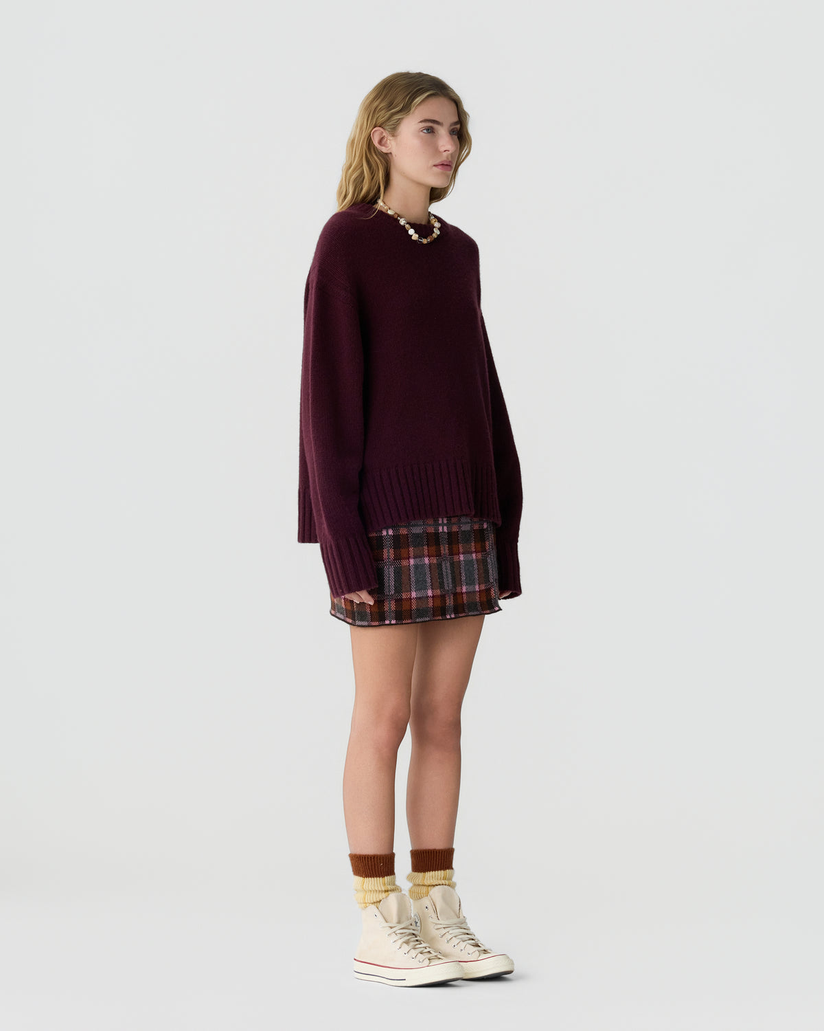 Woman wearing a burgundy oversized knit sweater paired with a plaid mini skirt, rust-colored socks, and off-white high-top sneakers, standing against a plain background.
