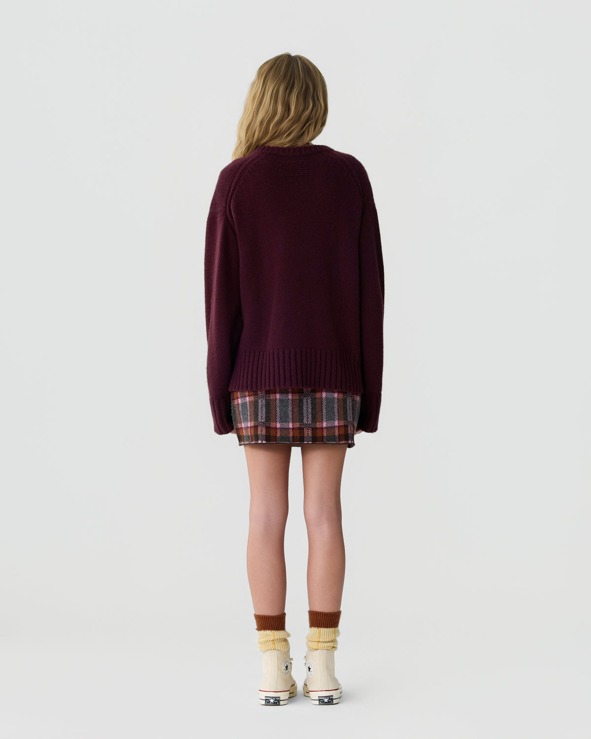 Woman standing facing away wearing a burgundy oversized sweater, a plaid mini skirt, slouched socks, and off-white sneakers against a plain light background.