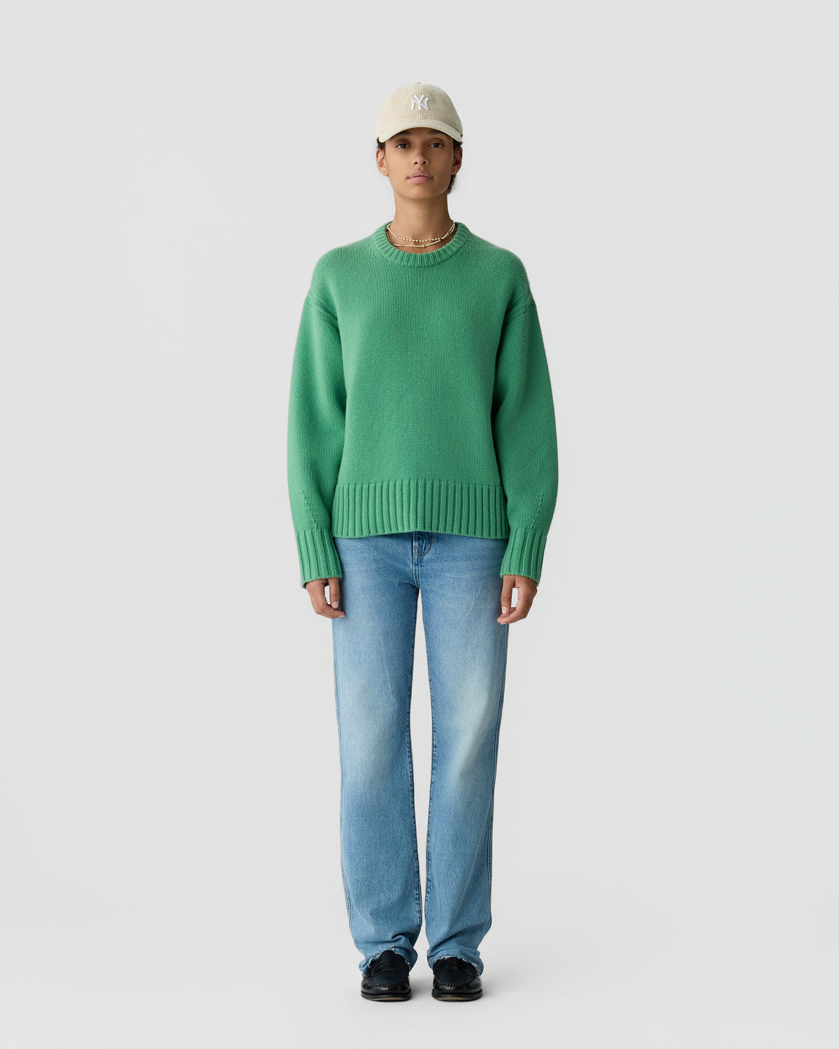 Person facing forward wearing a green crewneck sweater, light blue jeans, black shoes, and a beige baseball cap against a plain light gray background.