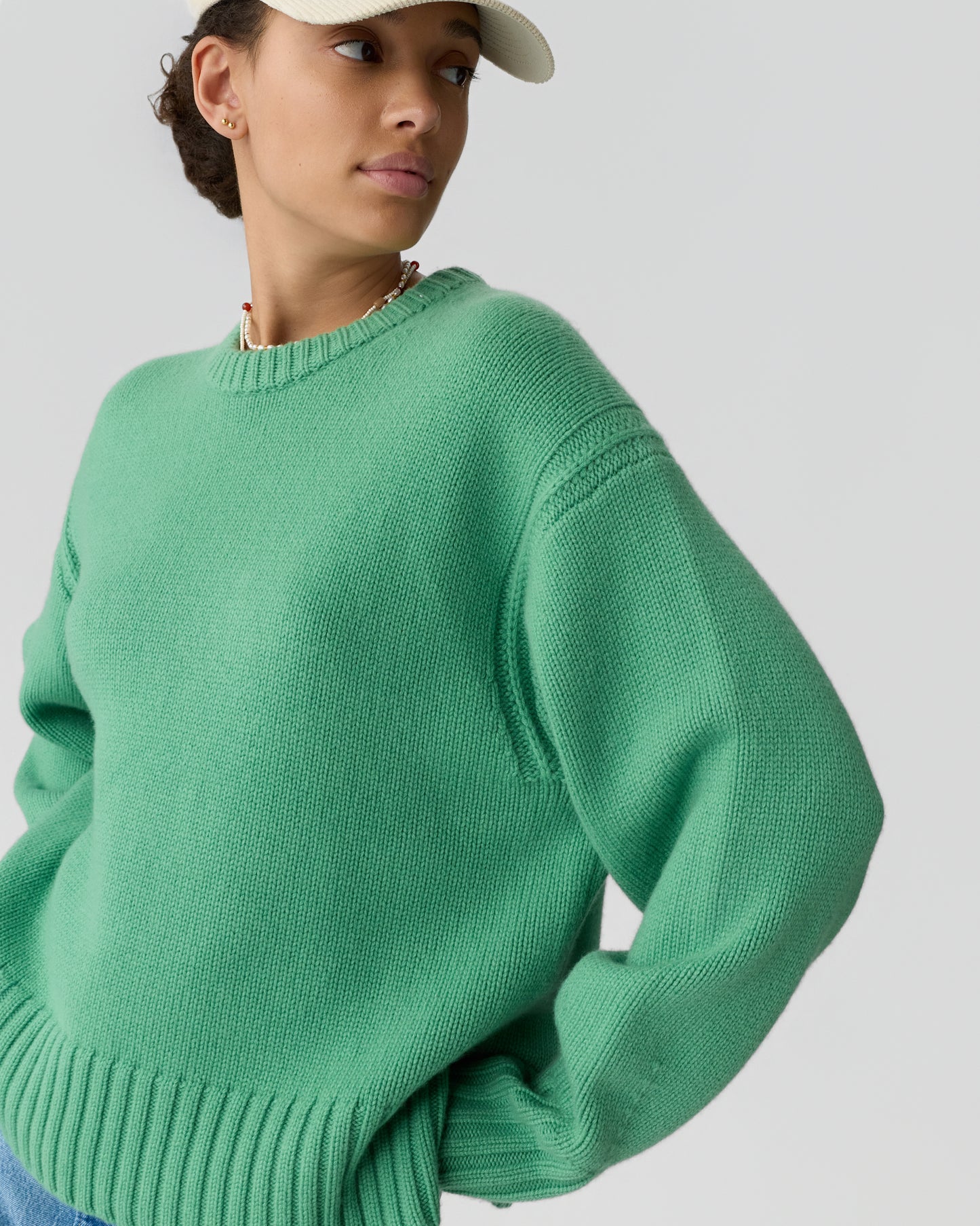 Woman wearing a green knitted sweater and a beige cap, looking to the side with hands in pockets against a plain light background.
