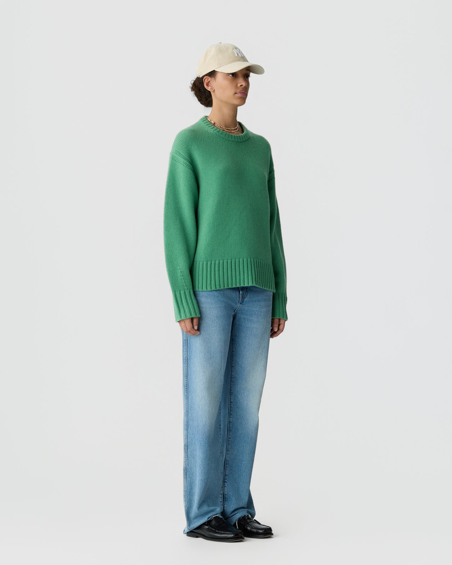 Person wearing a light beige baseball cap, a green long-sleeve cashmere sweater, blue jeans, and black shoes standing against a plain light background.