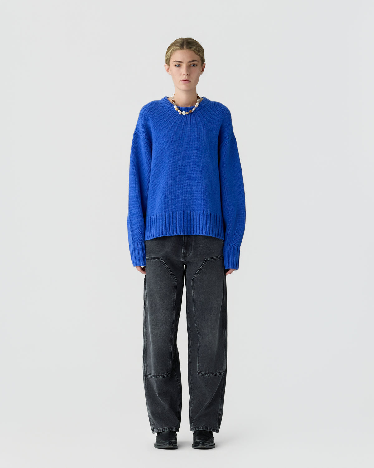 Person standing against a light gray background wearing a bright blue sweater with dark jeans and black shoes.