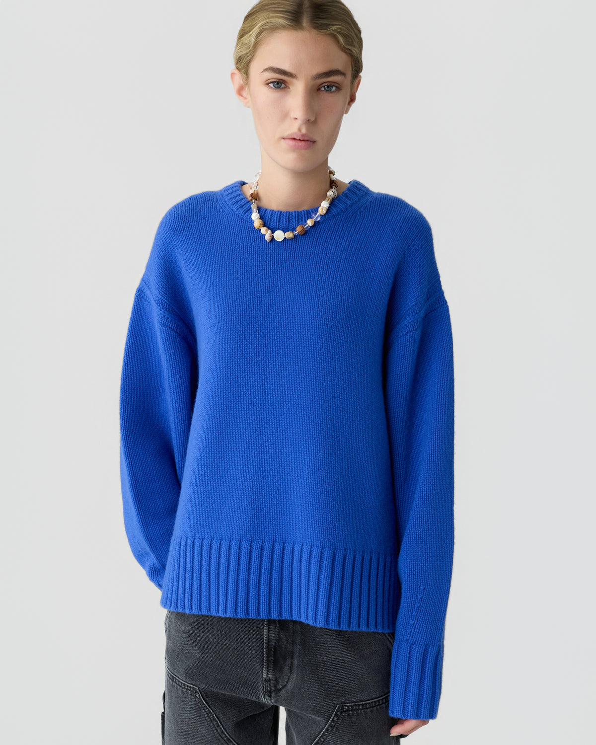 Model wearing a bright blue ribbed knit sweater with black jeans and a beaded necklace against a light gray background