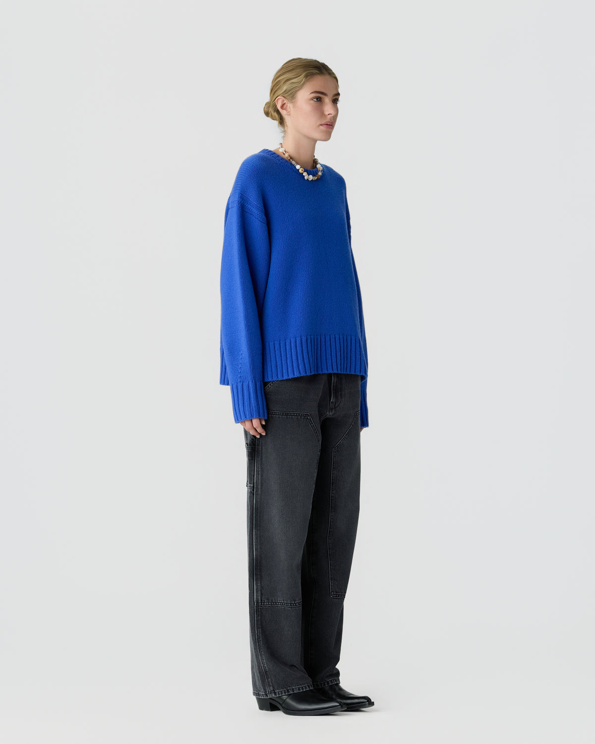 Model wearing a bright blue sweater with black jeans and black boots against a light gray studio background.