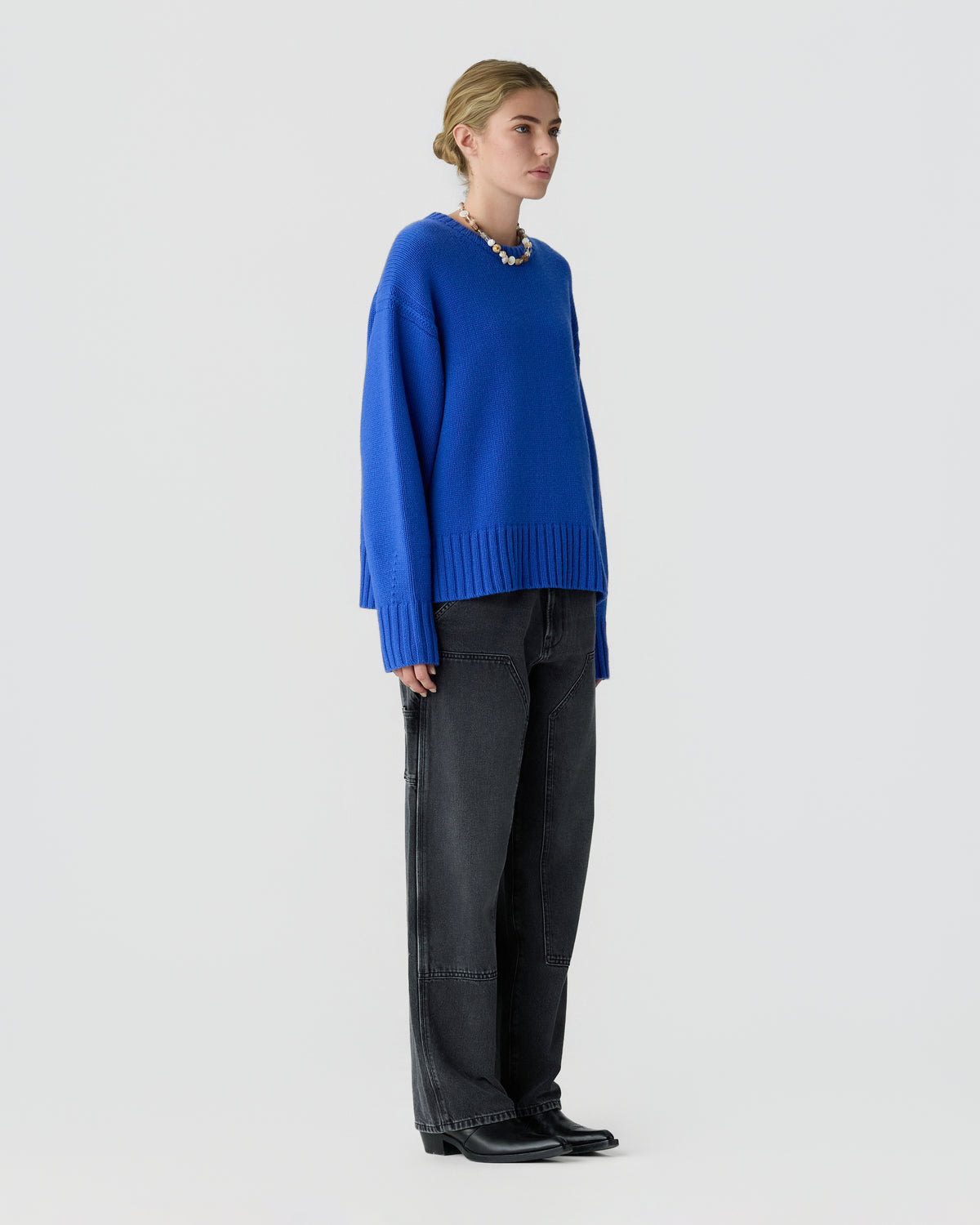 A model stands in profile wearing a bright blue cashmere sweater, dark wide leg jeans, black shoes, and a beaded necklace against a light gray background
