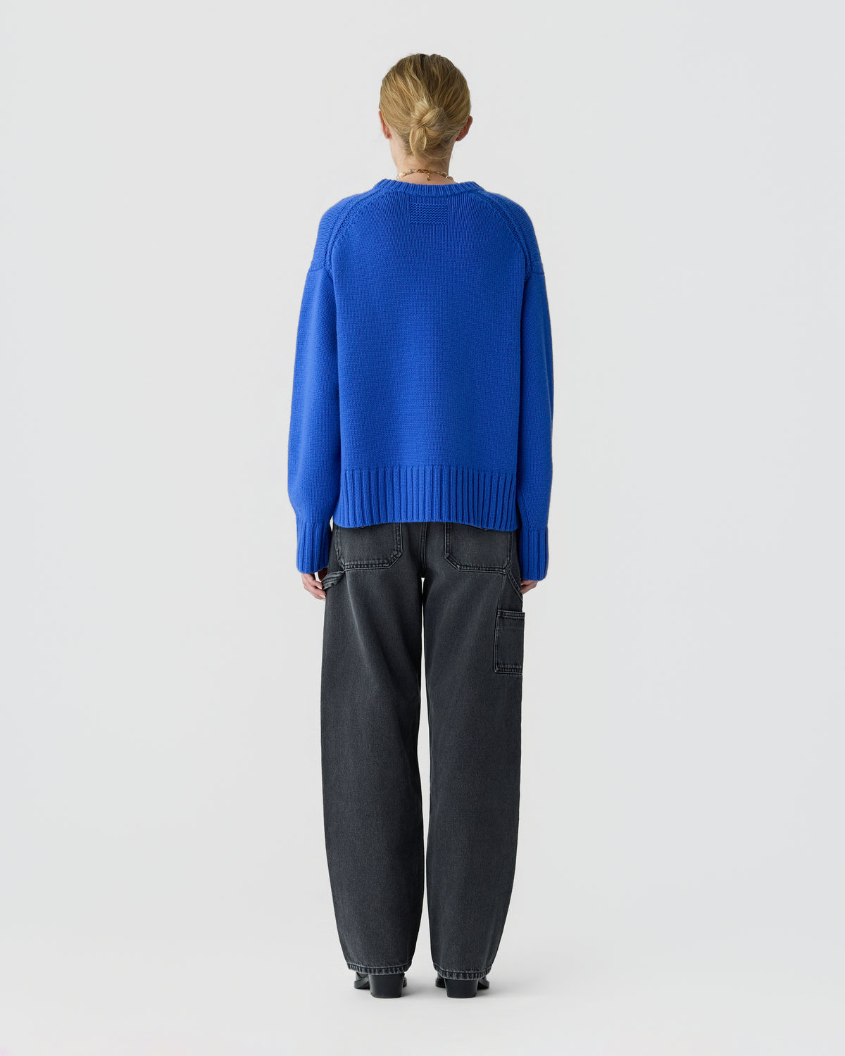 Person viewed from behind wearing a bright blue cashmere sweater with ribbed cuffs and hem, paired with relaxed black jeans and dark shoes against a pale background