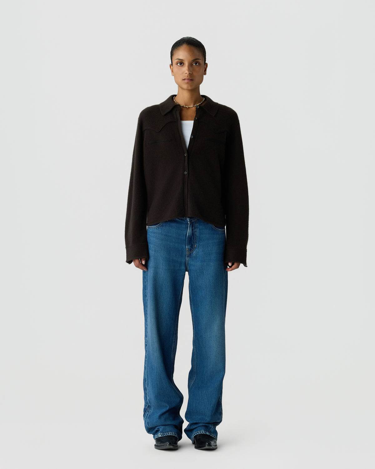 A woman stands facing forward wearing a black button-up long-sleeve shirt over a white top, paired with loose-fitting blue jeans and black shoes against a plain light gray background.