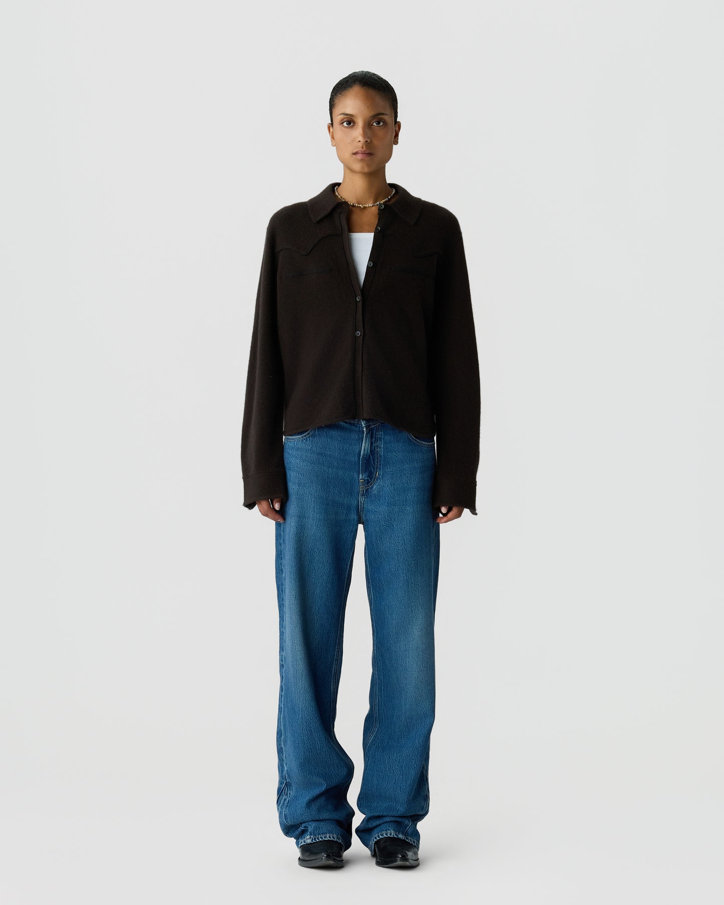A woman stands facing forward wearing a black button-up long-sleeve shirt over a white top, paired with loose-fitting blue jeans and black shoes against a plain light gray background.