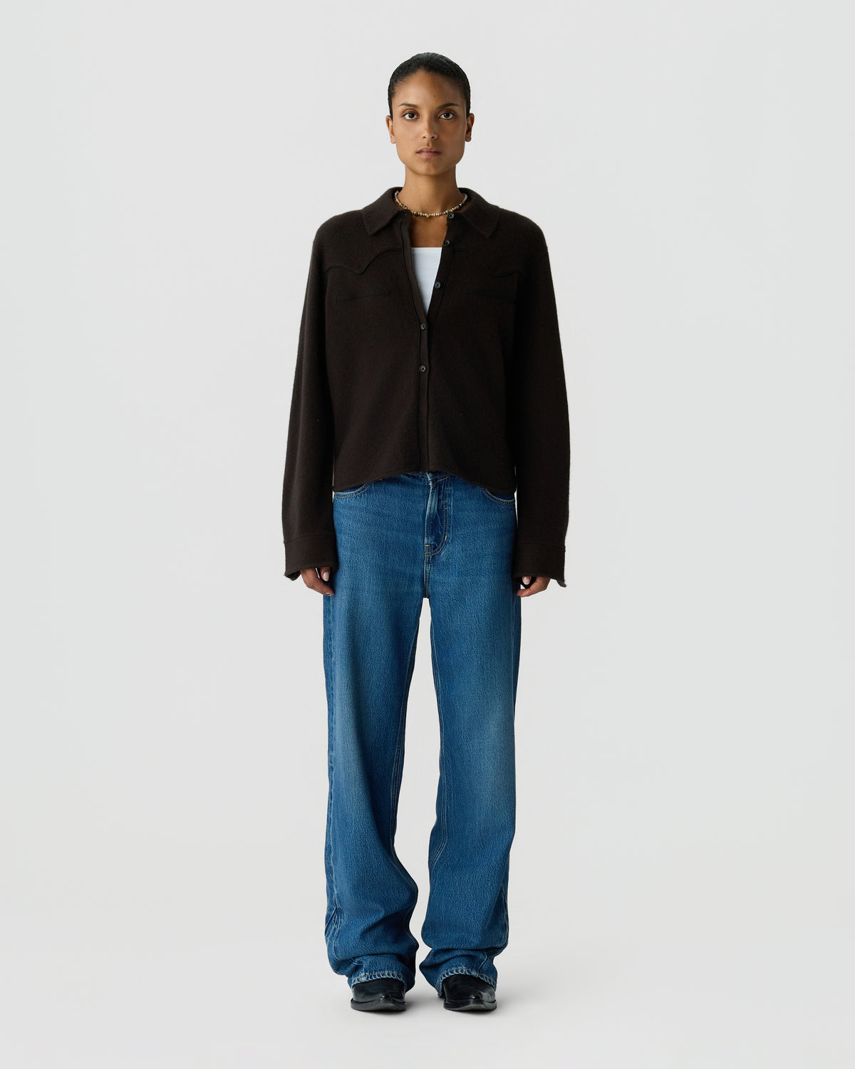Full-length portrait of a woman wearing a dark button-front jacket over a white top, relaxed blue jeans, and black shoes against a light background
