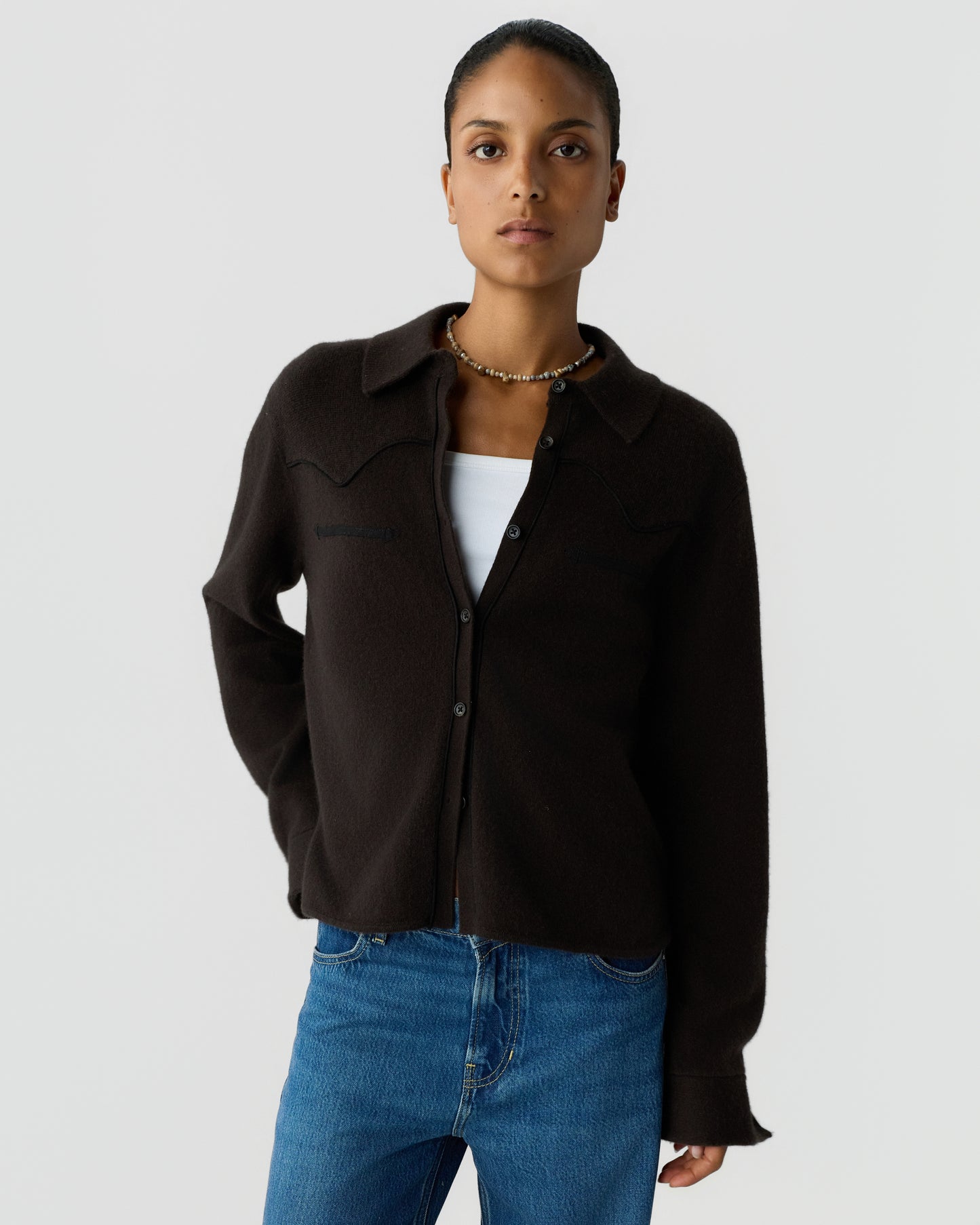 Young woman wearing a dark brown button-up cardigan over a white top and blue jeans, standing against a plain light gray background with a neutral expression.