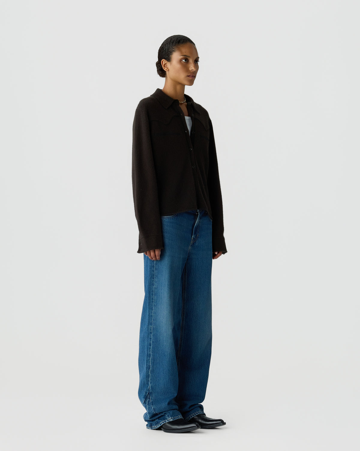 A woman with slicked-back hair stands wearing a black long-sleeve button-up shirt over a white top, paired with loose-fitting blue jeans and black shoes against a plain white background.