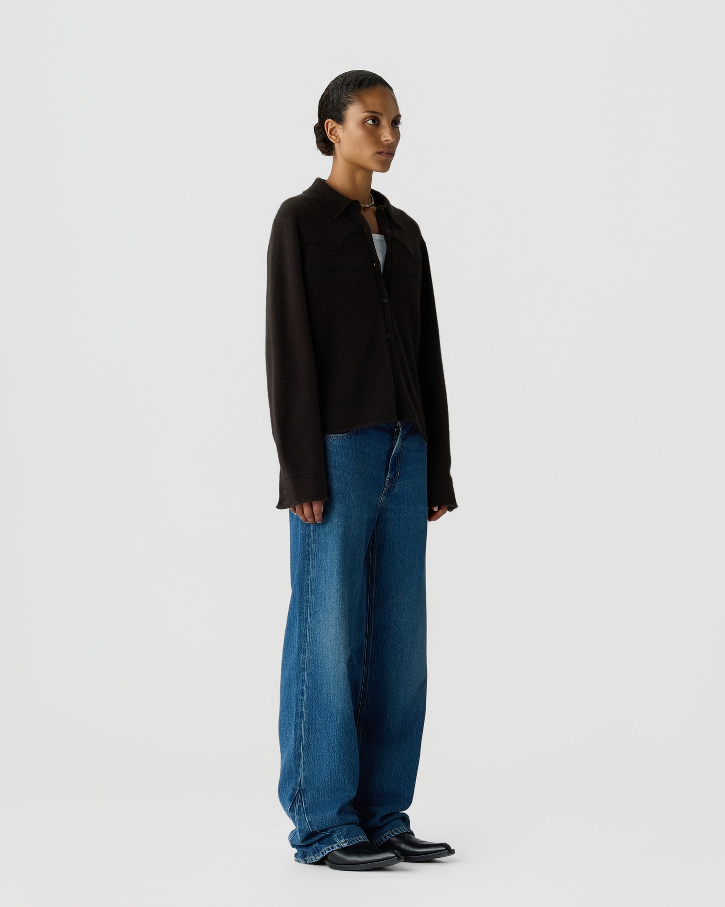 A woman with slicked-back hair stands wearing a black long-sleeve button-up shirt over a white top, paired with loose-fitting blue jeans and black shoes against a plain white background.