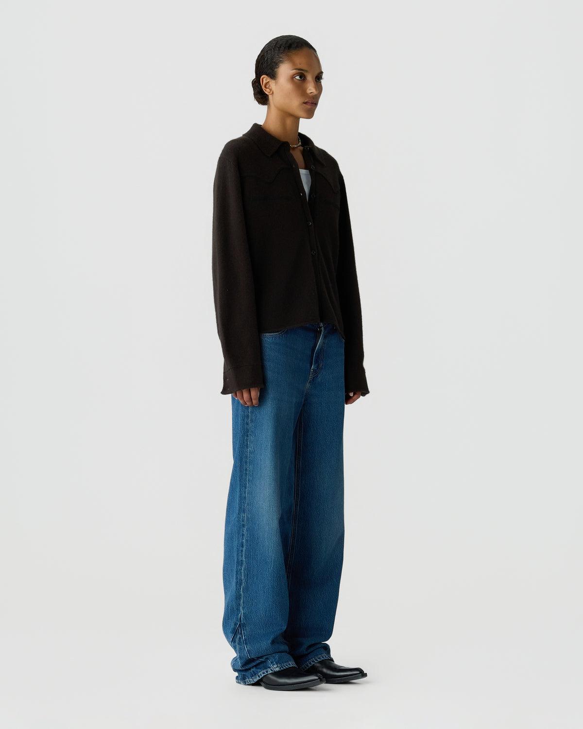 Full-length portrait of a woman wearing a dark long-sleeved jacket, loose blue jeans, and black shoes against a light gray background