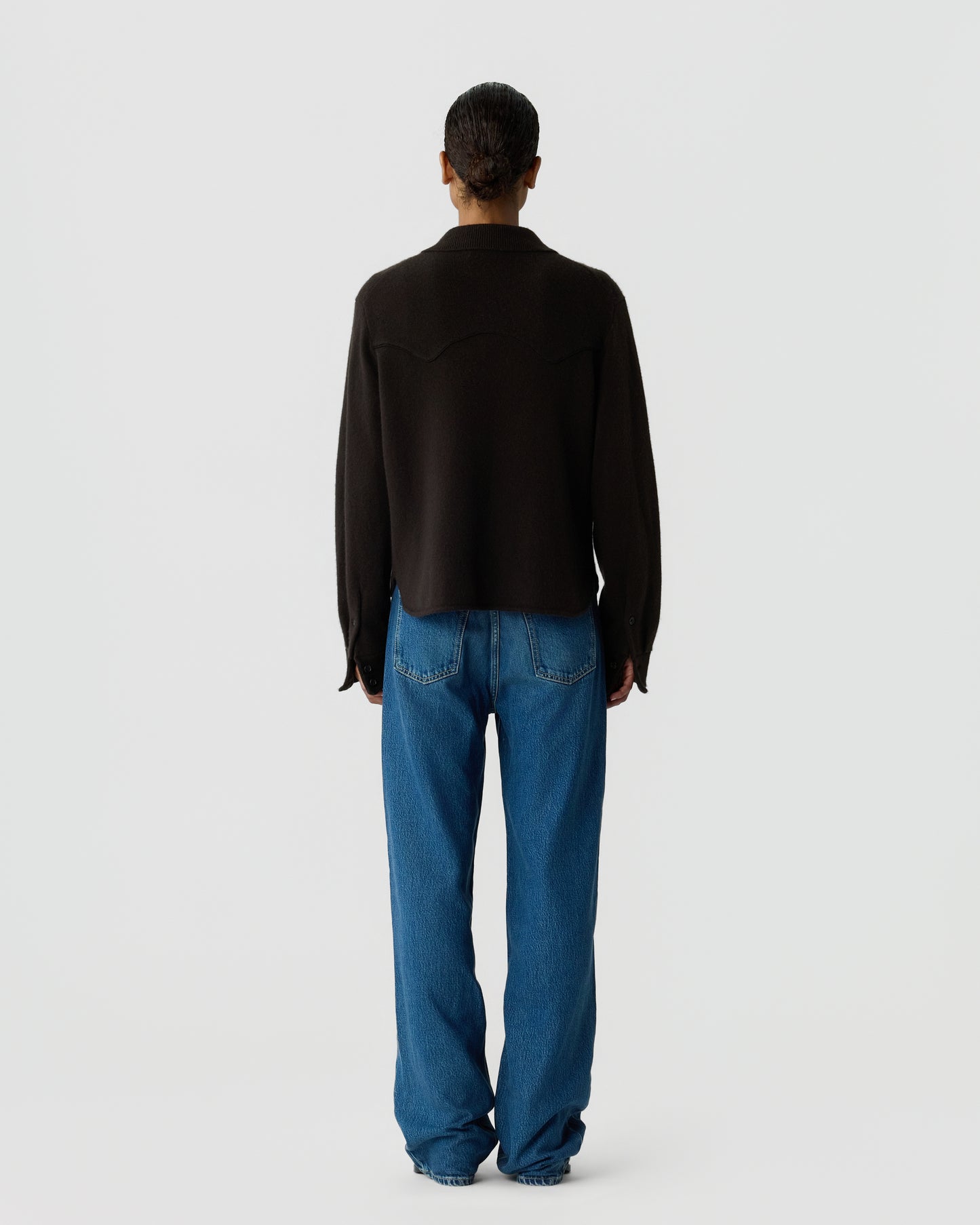 Person standing with back to camera wearing a black long-sleeve top and loose blue jeans against a plain light gray background.