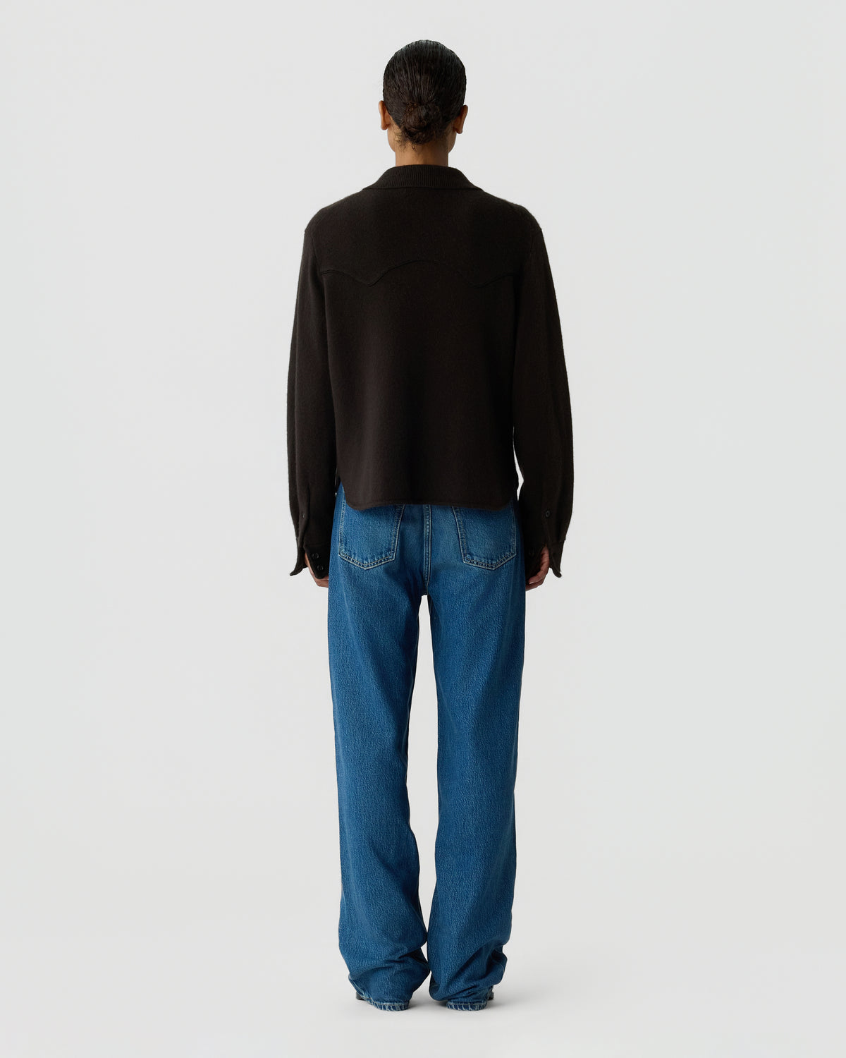 Back view of a person wearing a black long sleeve shirt with blue jeans against a plain light gray background