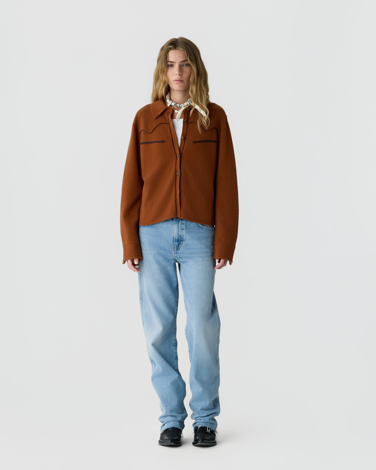 A woman stands facing forward wearing a rust-colored button-up jacket over a white top, light blue jeans, black shoes, and a white patterned scarf tied around her neck against a plain gray background.