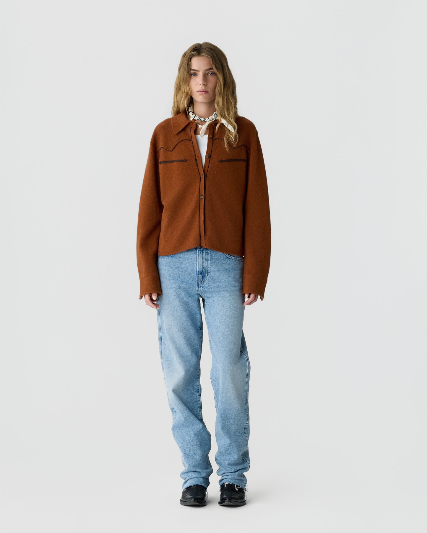 A woman stands facing forward wearing a rust-colored button-up jacket over a white top, light blue jeans, black shoes, and a white patterned scarf tied around her neck against a plain gray background.