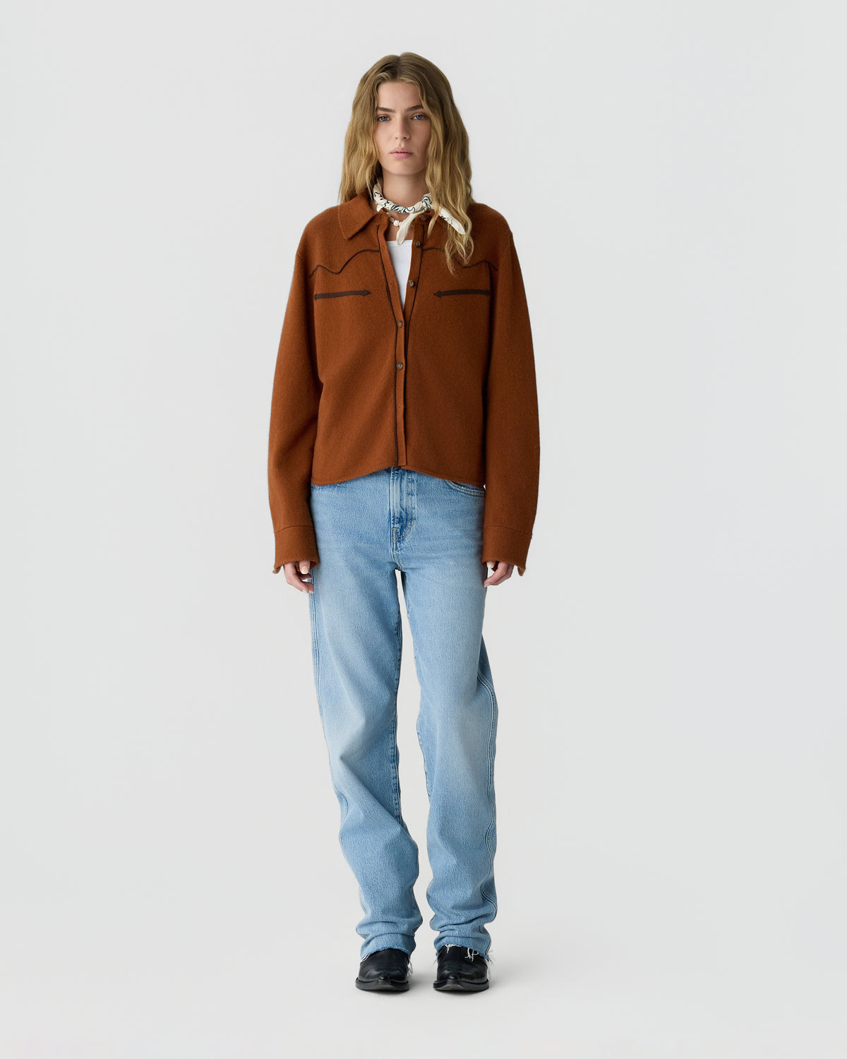 Full-length portrait of a woman wearing a rust brown button-front jacket over a white top, light blue jeans, and black shoes against a light gray background