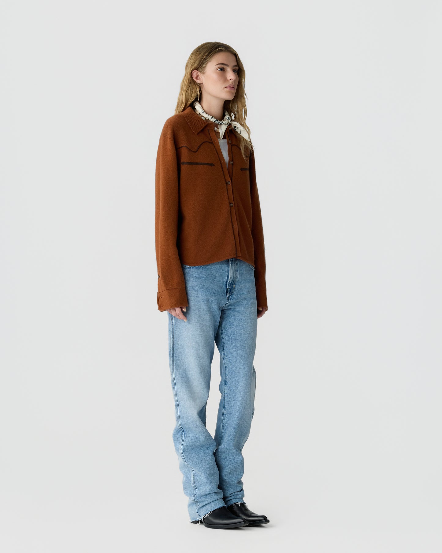 Woman wearing a brown button-up cardigan, light blue jeans, black shoes, and a patterned scarf tied around her neck, standing against a plain white background.