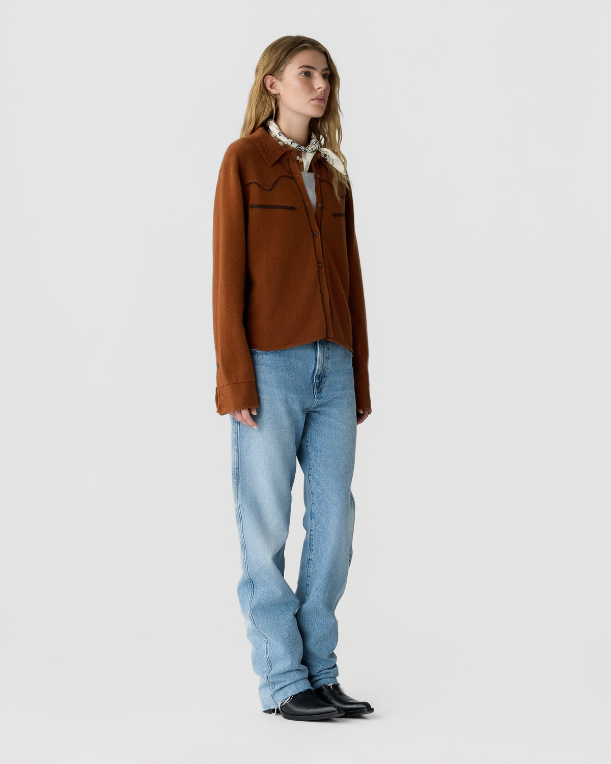 Woman standing in a brown zip-front sweater, light blue jeans, and black shoes against a plain light gray background